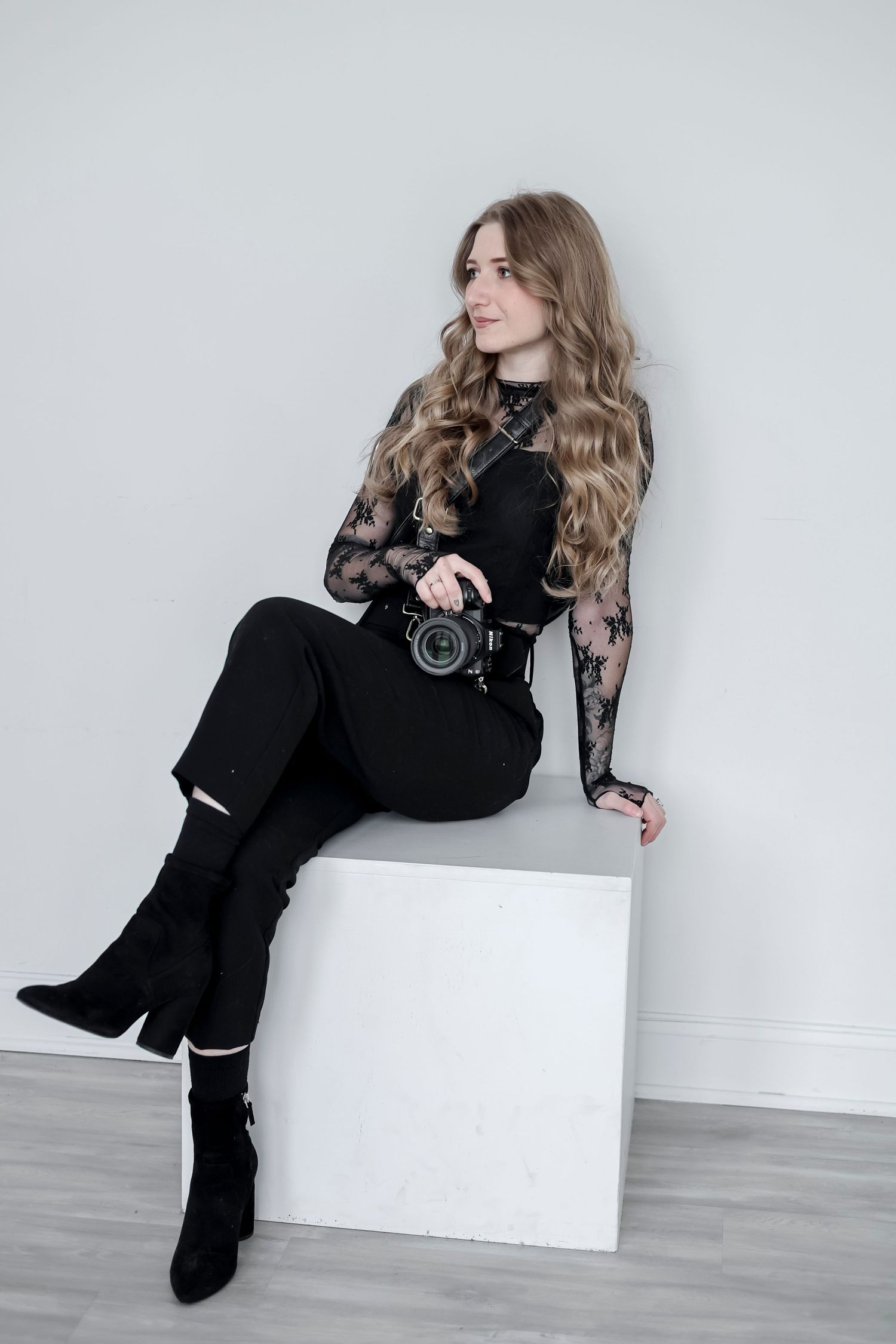 Model in black outfit poses elegantly against white wall in fashion photoshoot series.