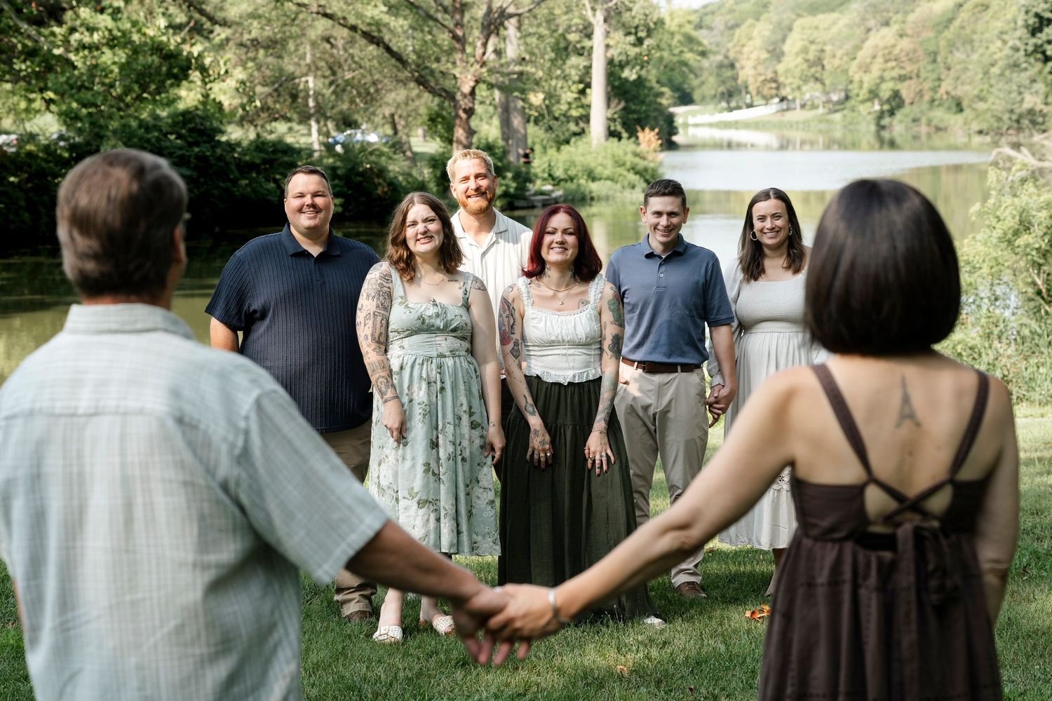 Group of friends holding hands outdoors near a lake, smiling and posing for a family photo in a lush green setting.