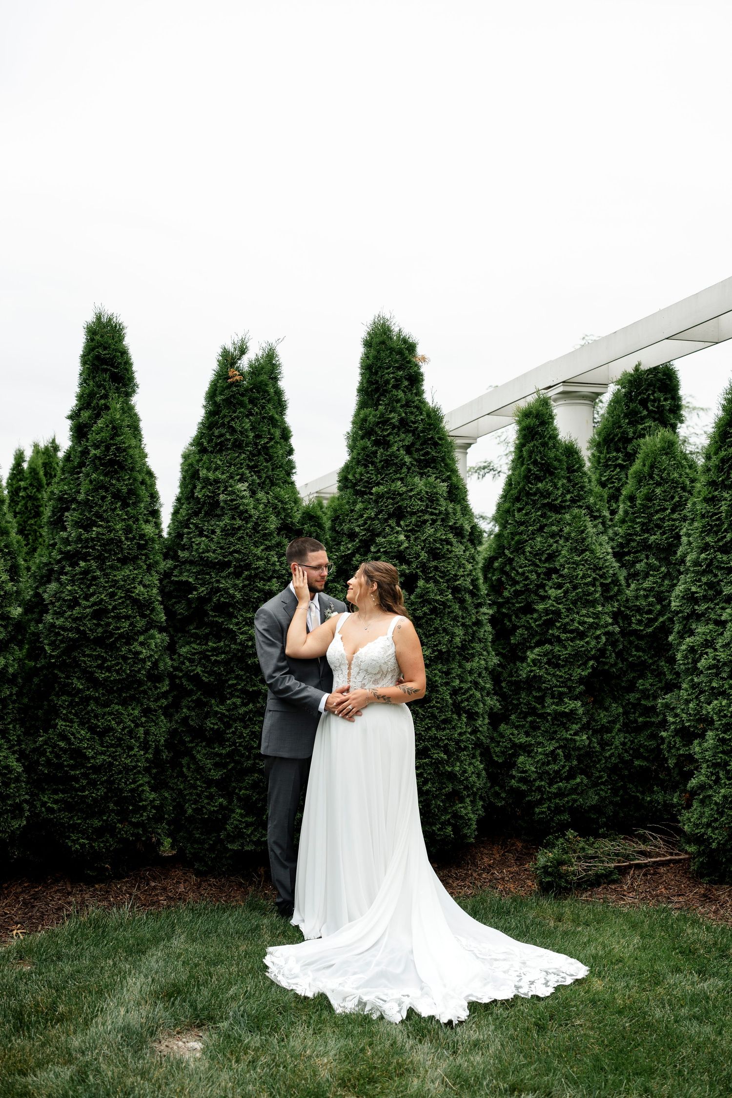 Bride and groom share a romantic moment in front of tall evergreen trees on their wedding day.