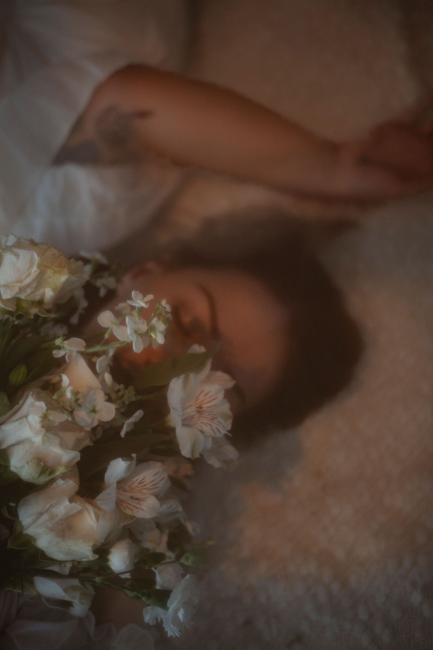 Woman lying down with white flowers in foreground, soft dreamy bokeh photography with muted tones.