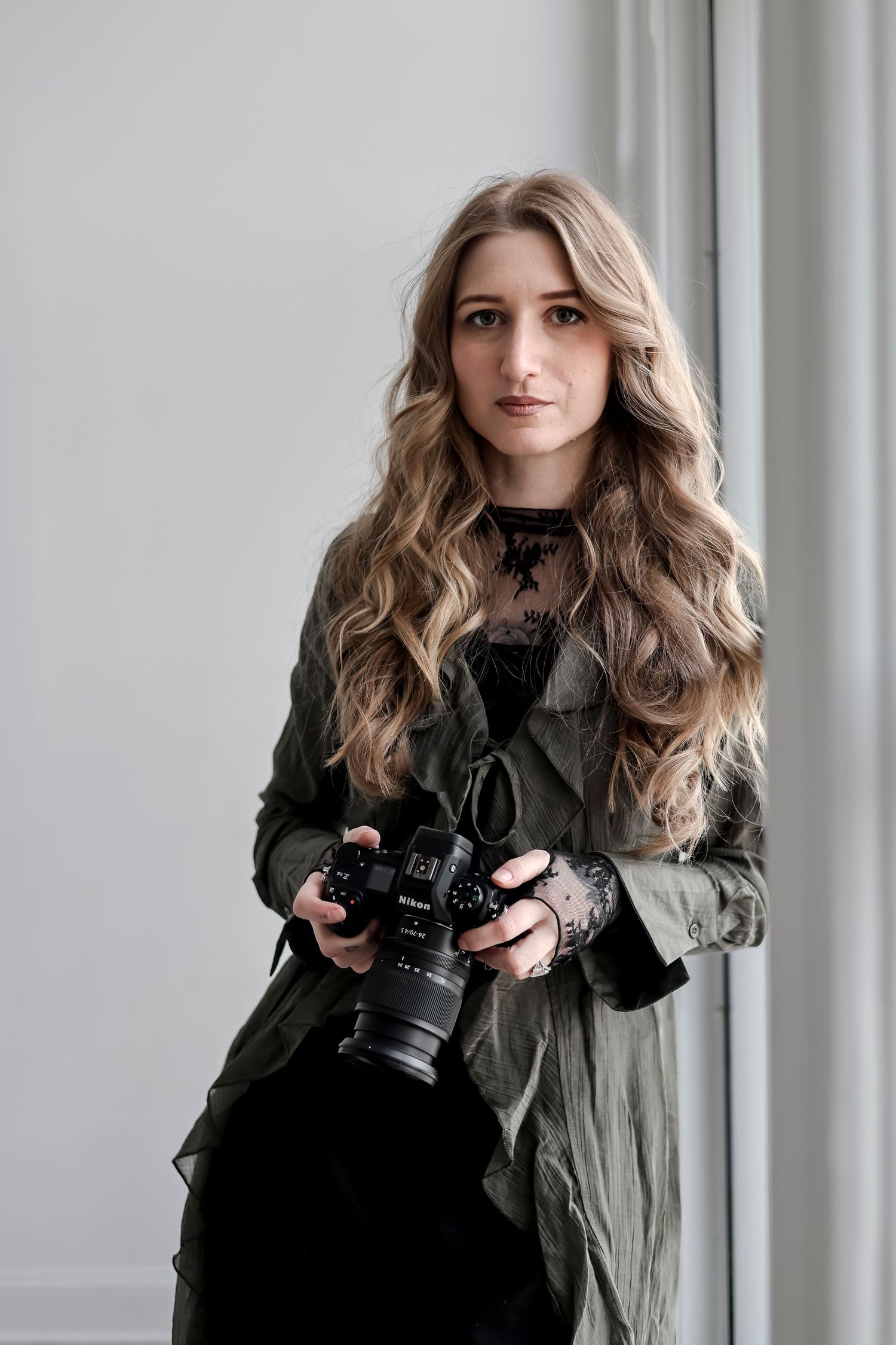 Professional photographer wearing olive green jacket and black outfit holds DSLR camera against light gray background.
