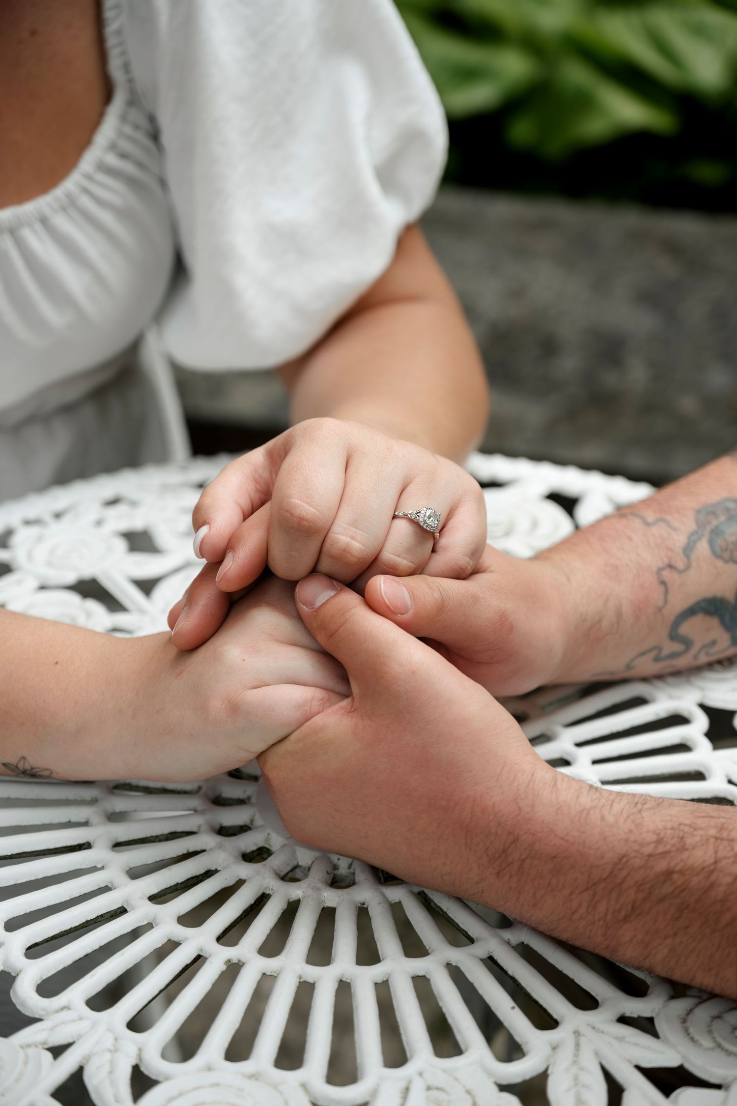 Couple holding hands at white ornate table, woman wearing diamond engagement ring, man with tattoo visible.