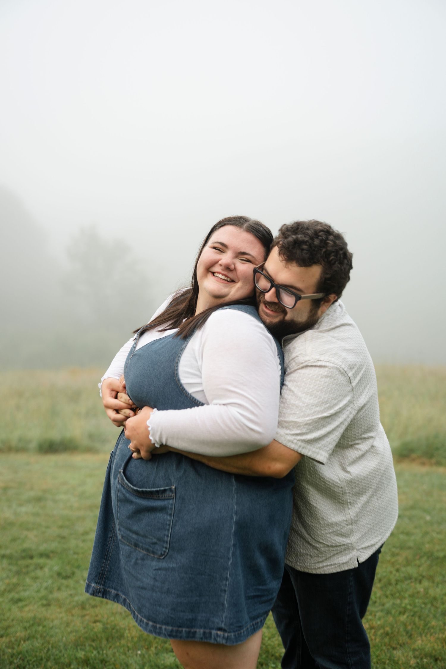 A joyful couple embraces in a misty field, woman in denim dress laughing as man hugs her from behind.