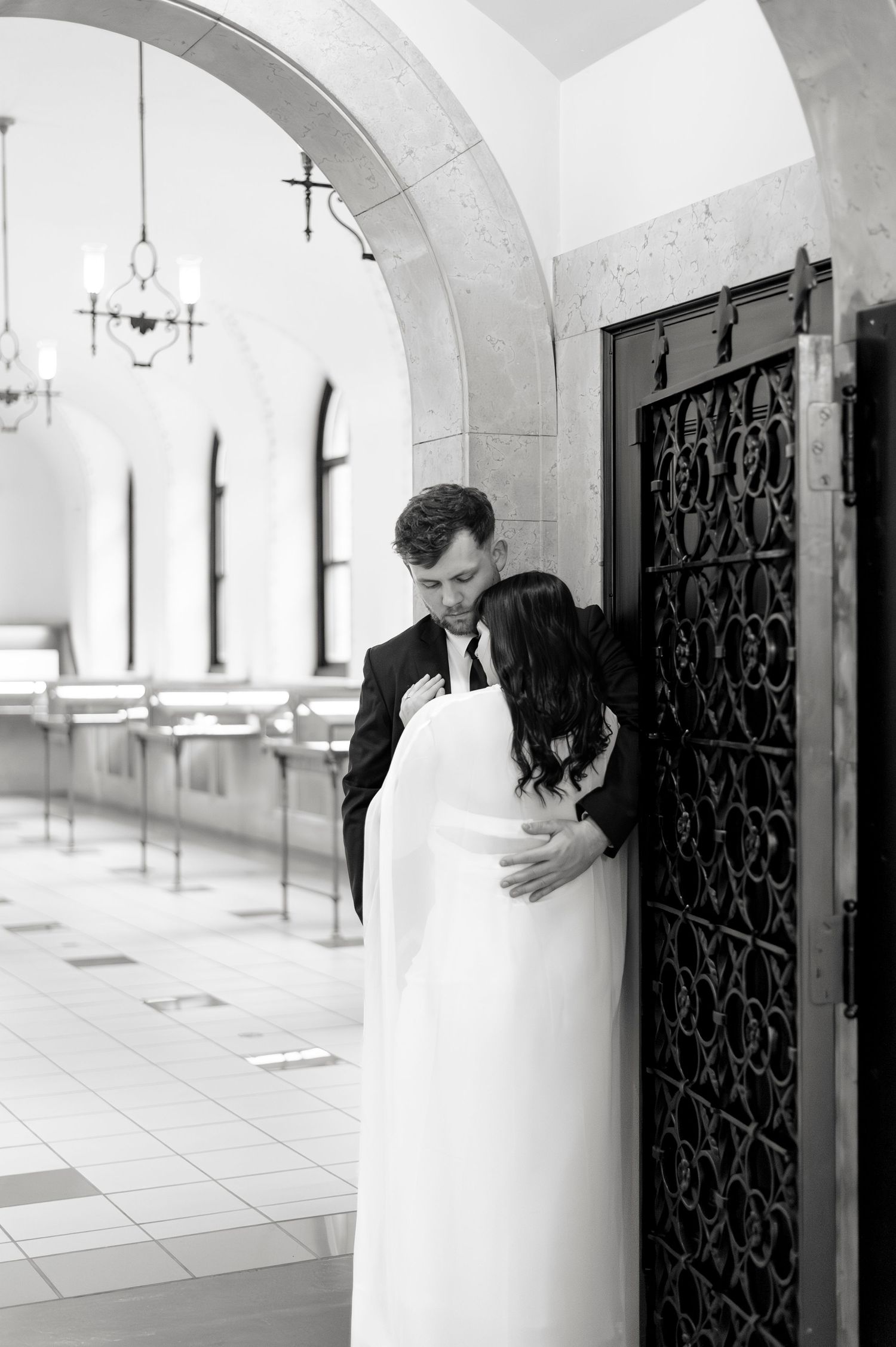 Black and white photo of groom embracing bride beside ornate iron door in arched stone hallway.