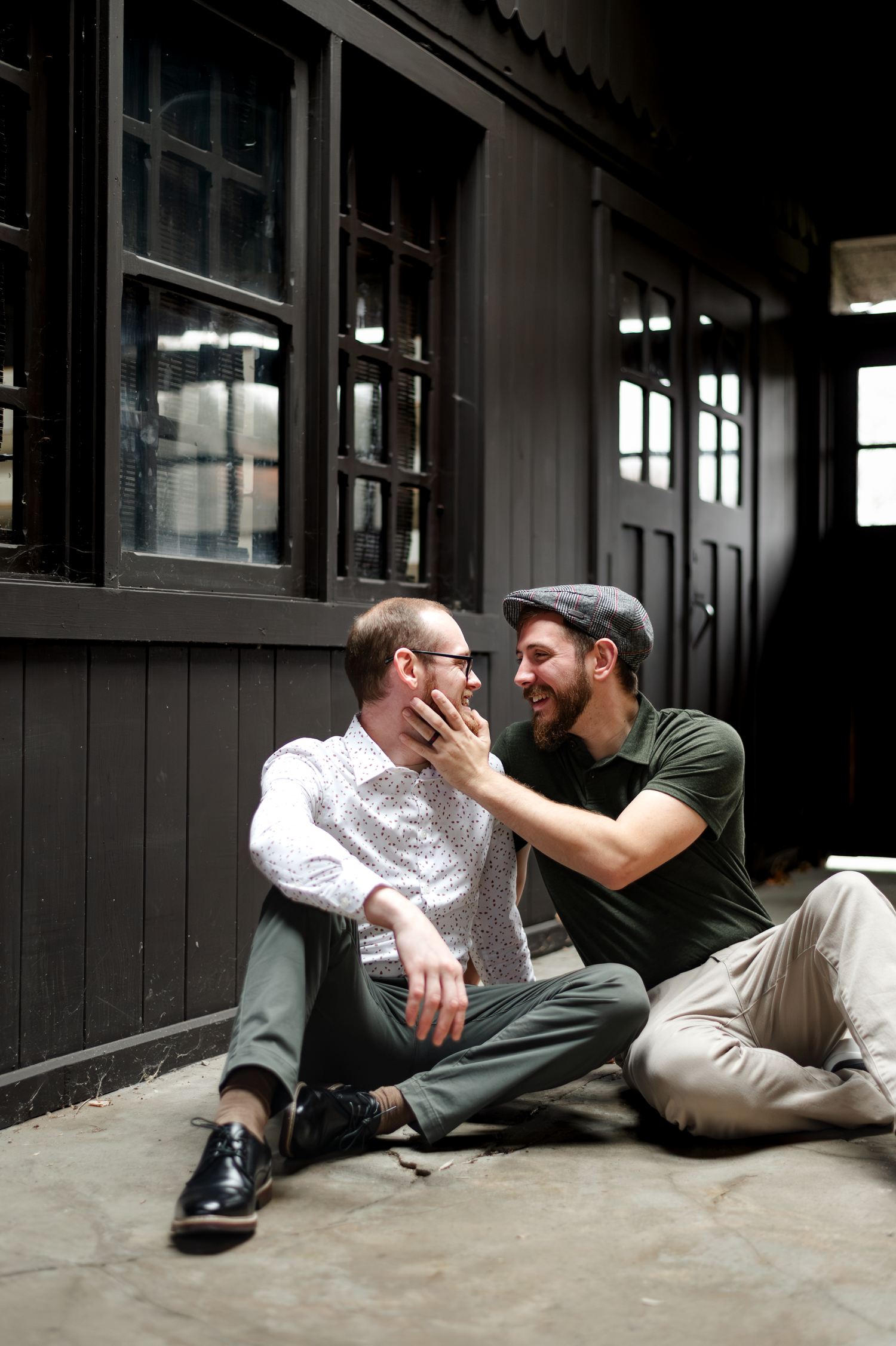 Two men sitting on the ground outside a dark building, sharing a tender moment as one gently touches the others face.