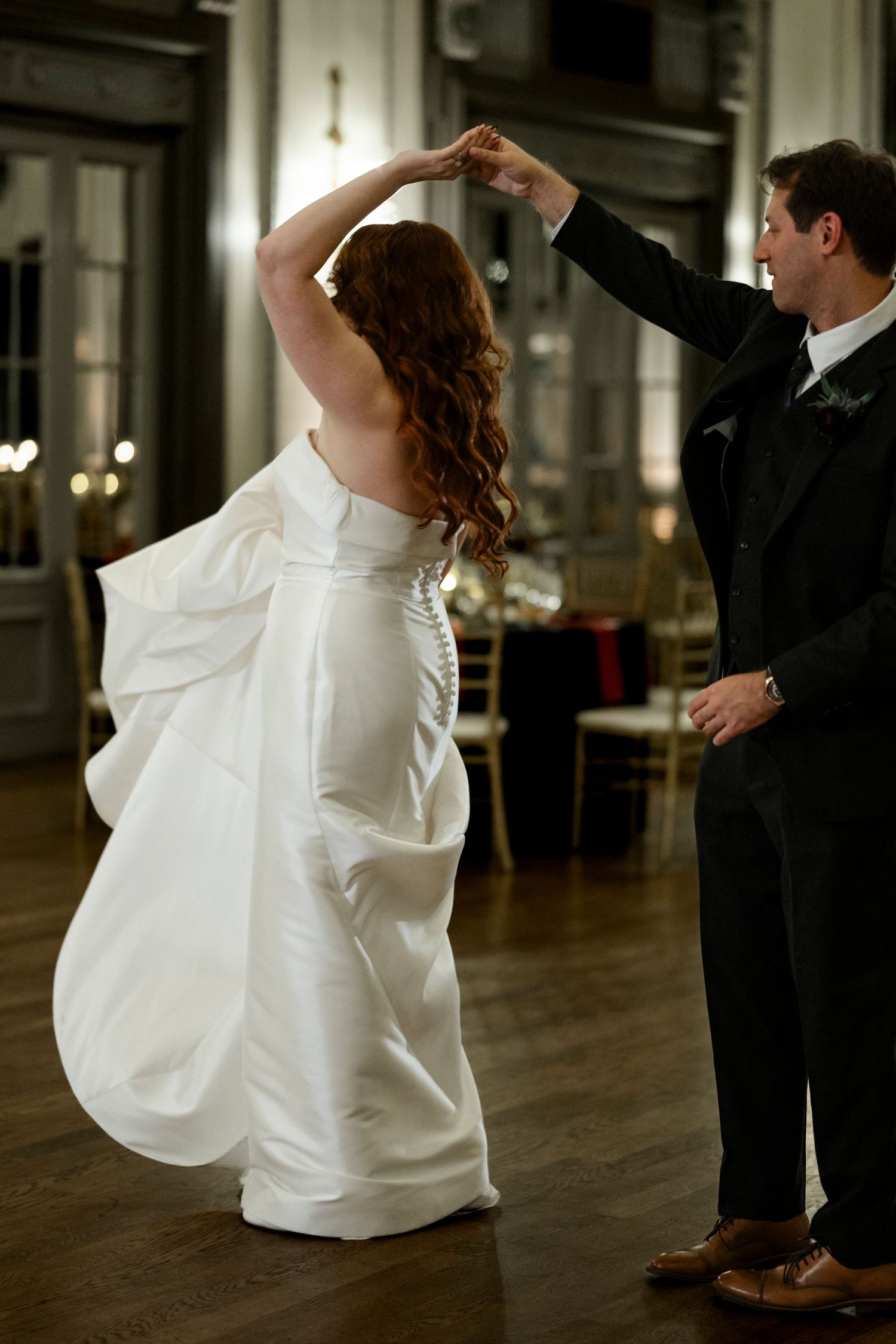 A bride and groom dance together in an elegant ballroom with dramatic lighting and wooden floors.