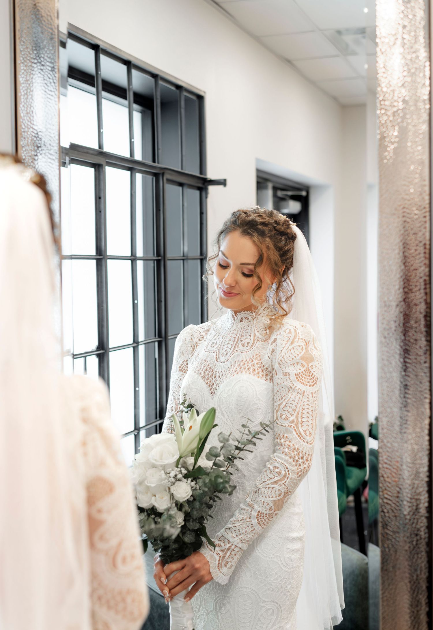 A woman wearing a long, white lace wedding dress stands in a hallway holding a bouquet of white flowers.