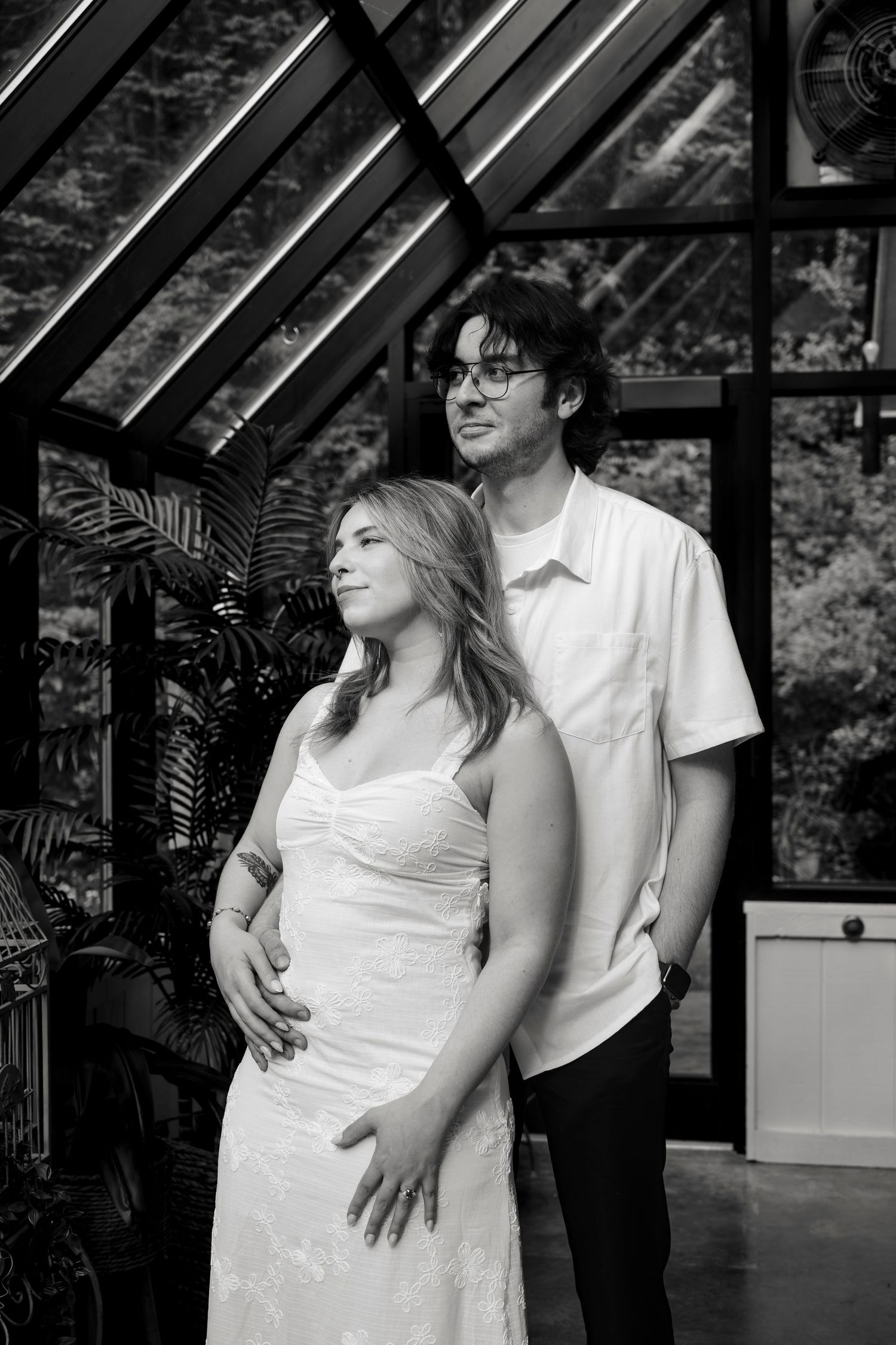 Black and white photo of a pregnant couple posing in a greenhouse, man embracing woman from behind.