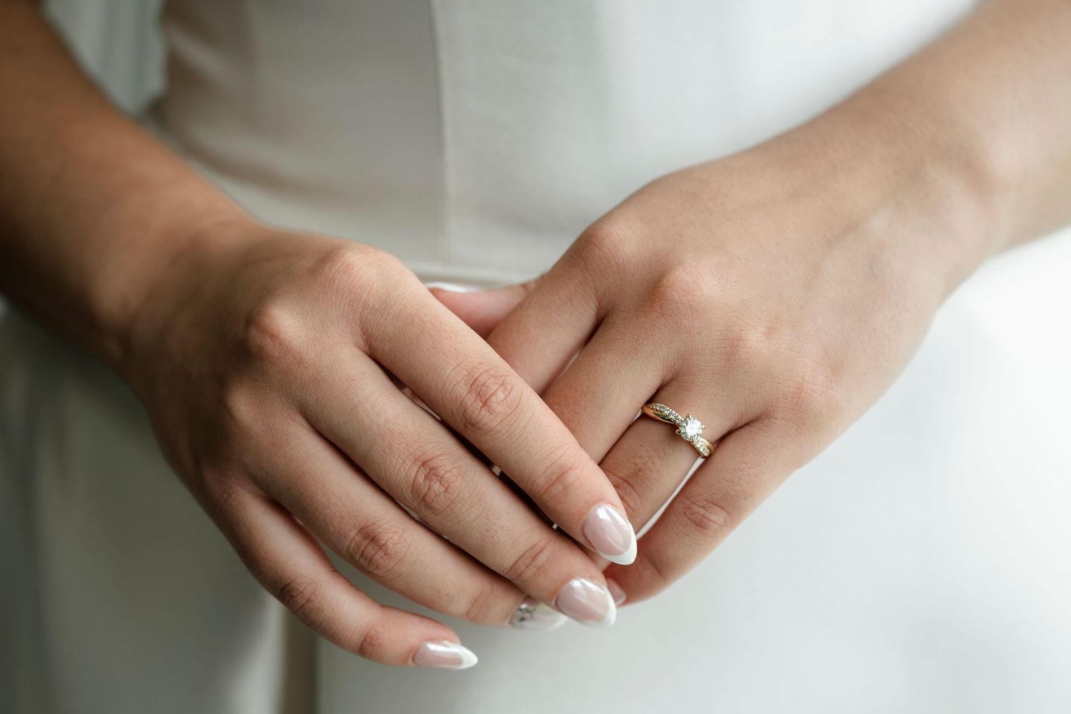 Close-up of bride's hands with elegant diamond engagement ring and wedding band on white dress.