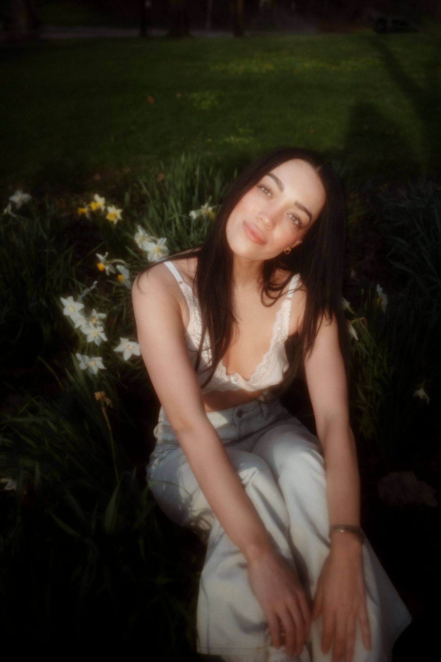 Young woman in white lace top and jeans sitting among white daffodils in sunny garden.