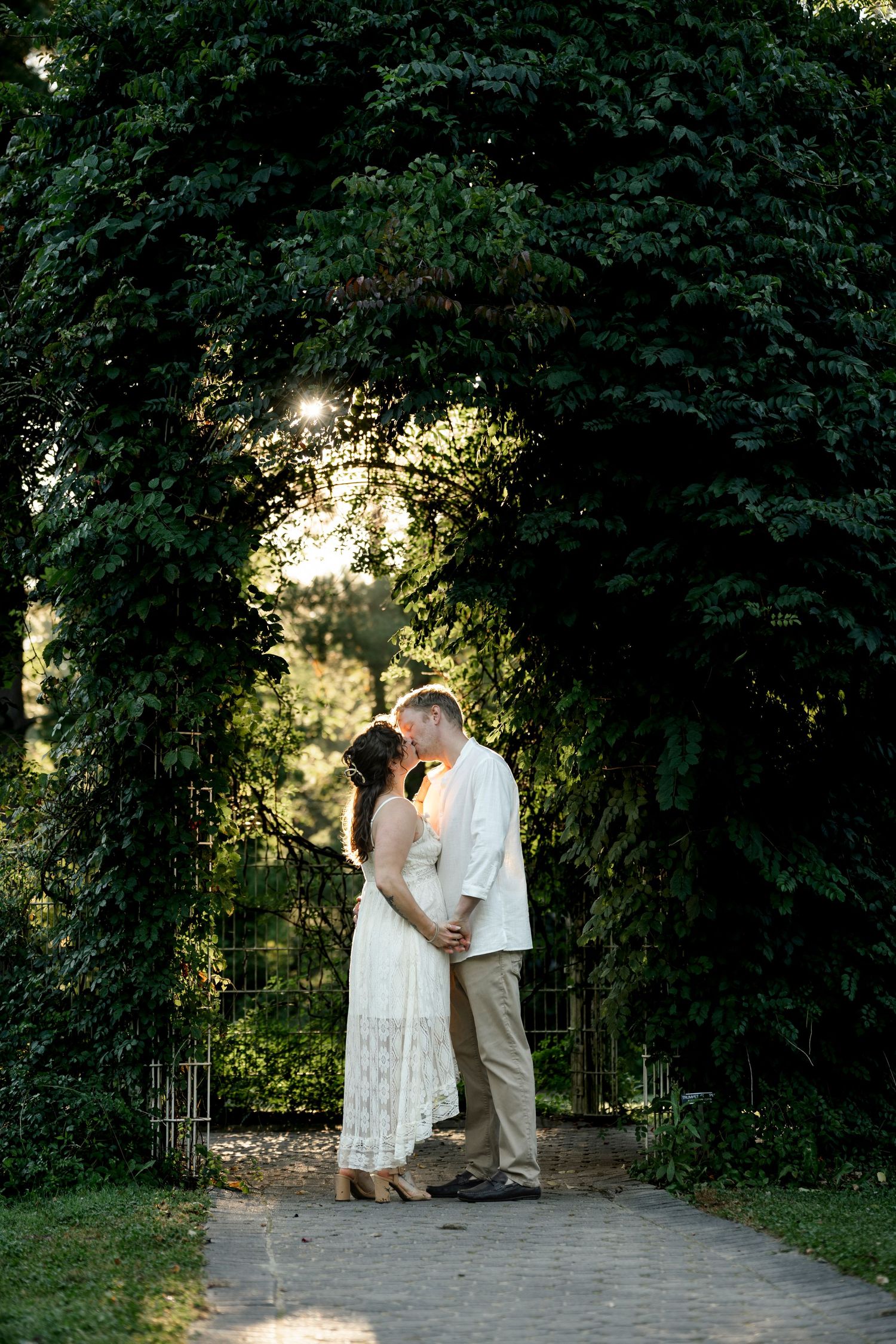 Couple sharing a romantic kiss under a lush green garden archway in golden hour light, bride in white lace dress.