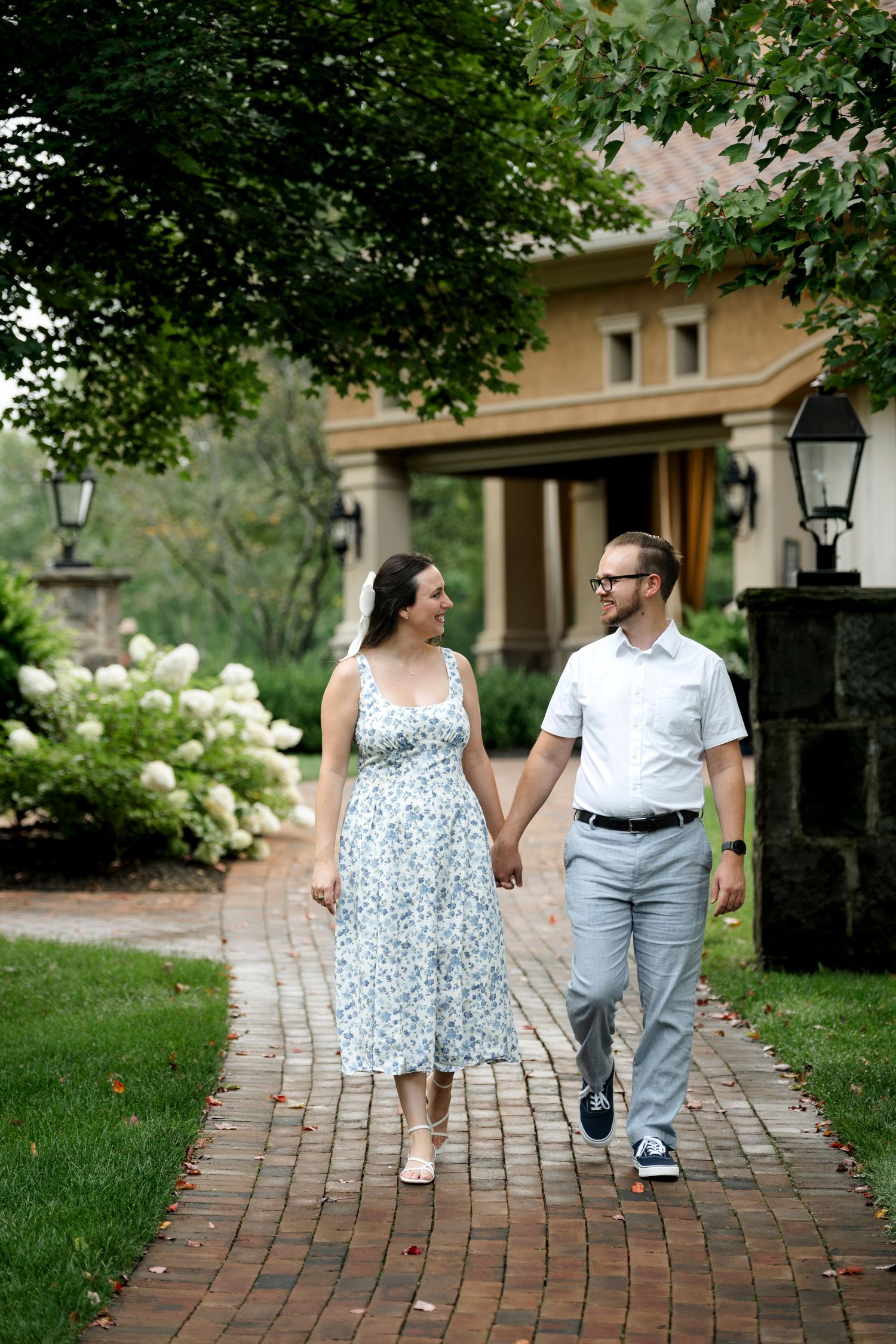 Couple holding hands walking on brick path past blooming hydrangeas outside elegant home.