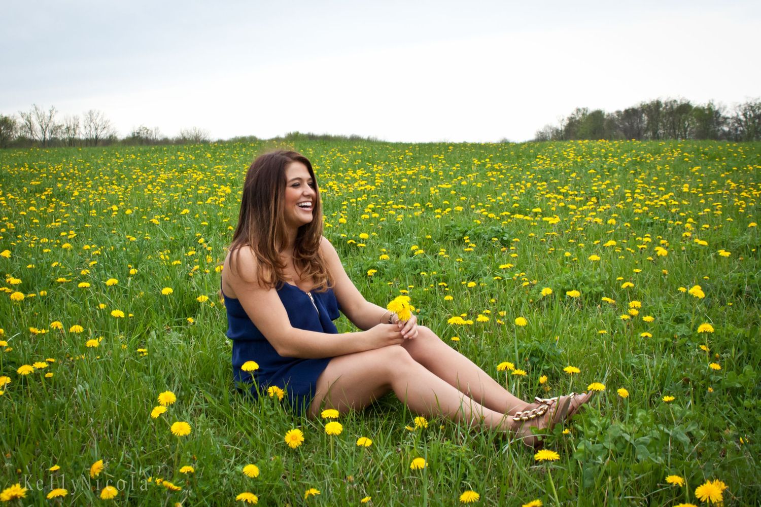 Someone in a blue dress sits in a vibrant field of yellow dandelions on a spring day.