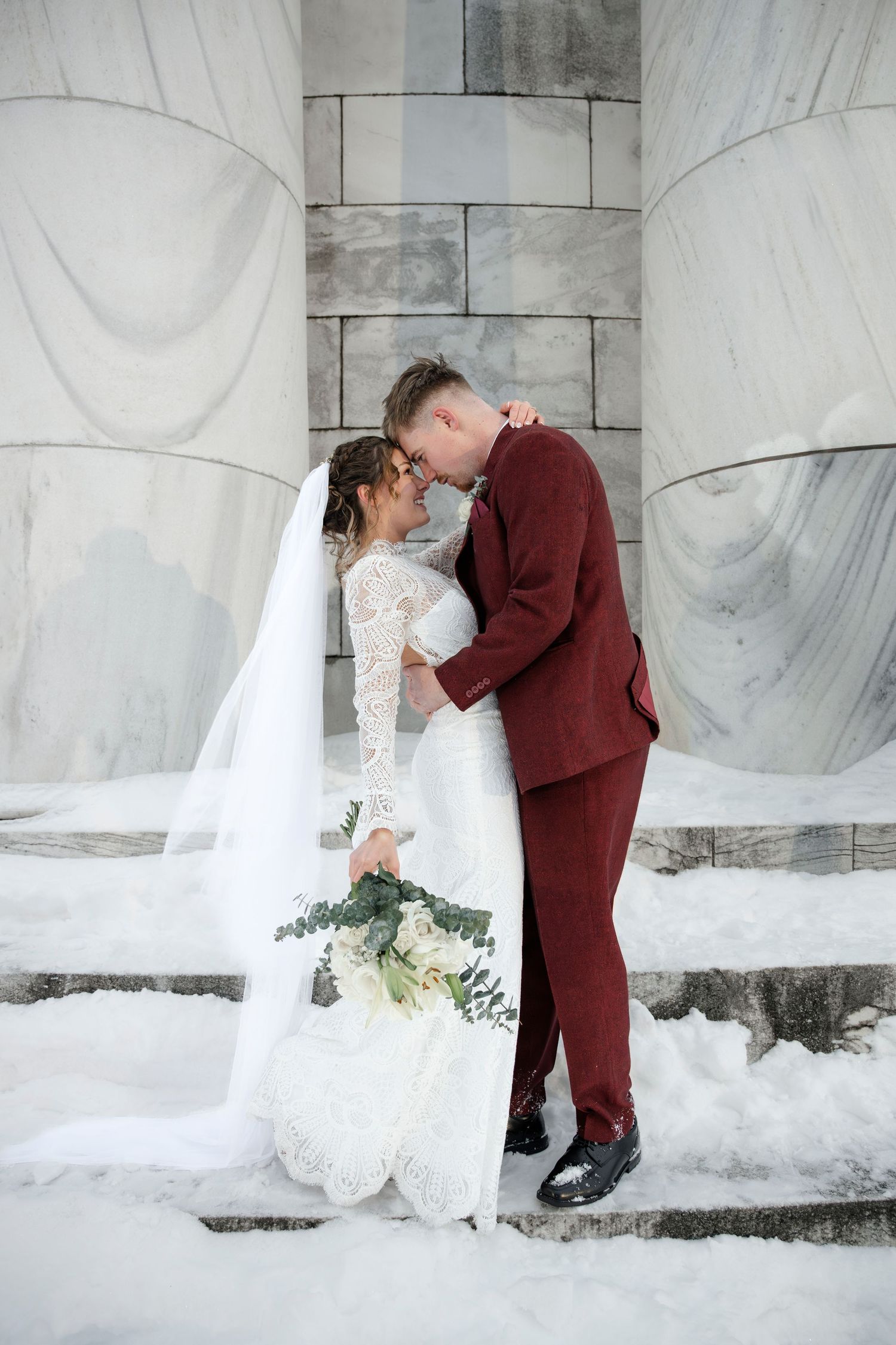 A bride and groom embrace on a snowy staircase, dressed in a white wedding gown and a burgundy suit.