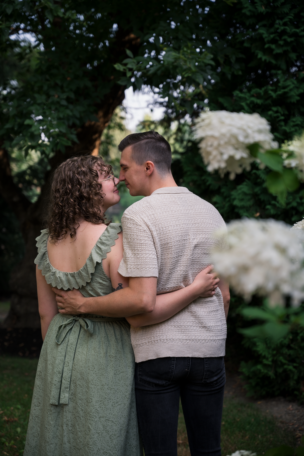 Couple embracing in a lush garden with white hydrangeas, wearing green dress and beige knit shirt.