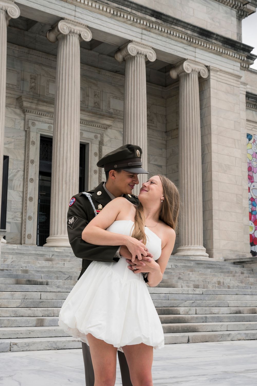 A soldier in uniform embraces a woman in a white dress on steps of a neoclassical building with tall columns.