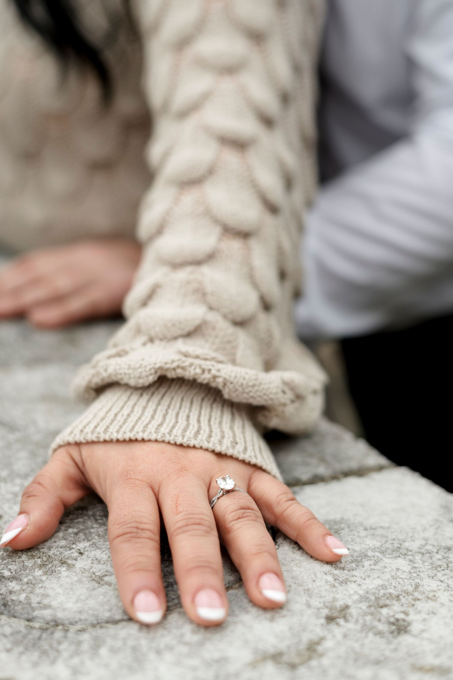 Close-up of woman's hand displaying diamond engagement ring, wearing beige cable-knit sweater sleeve on stone surface.
