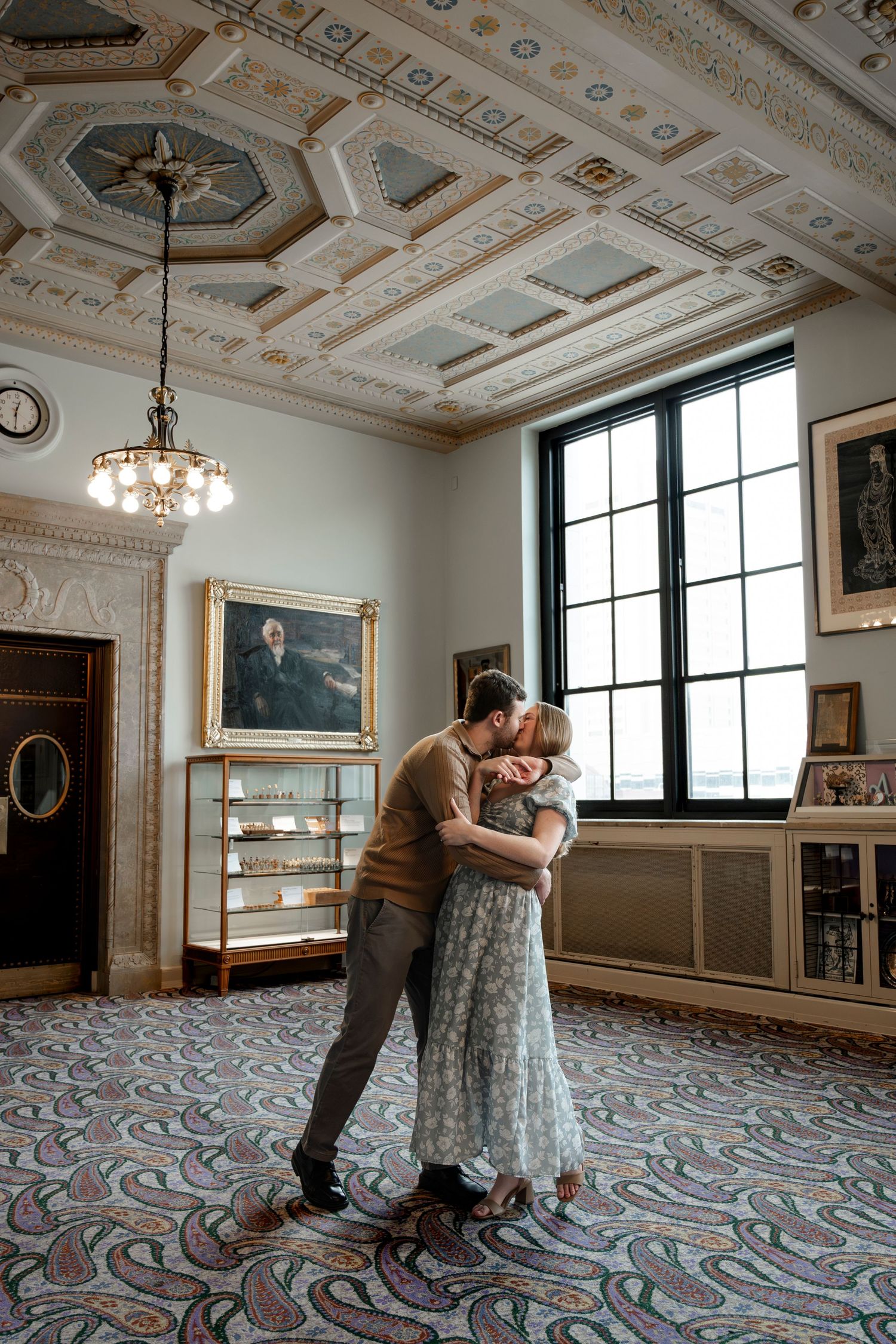 Couple sharing a romantic embrace in an elegant room with ornate ceiling, paisley carpet, and large windows.