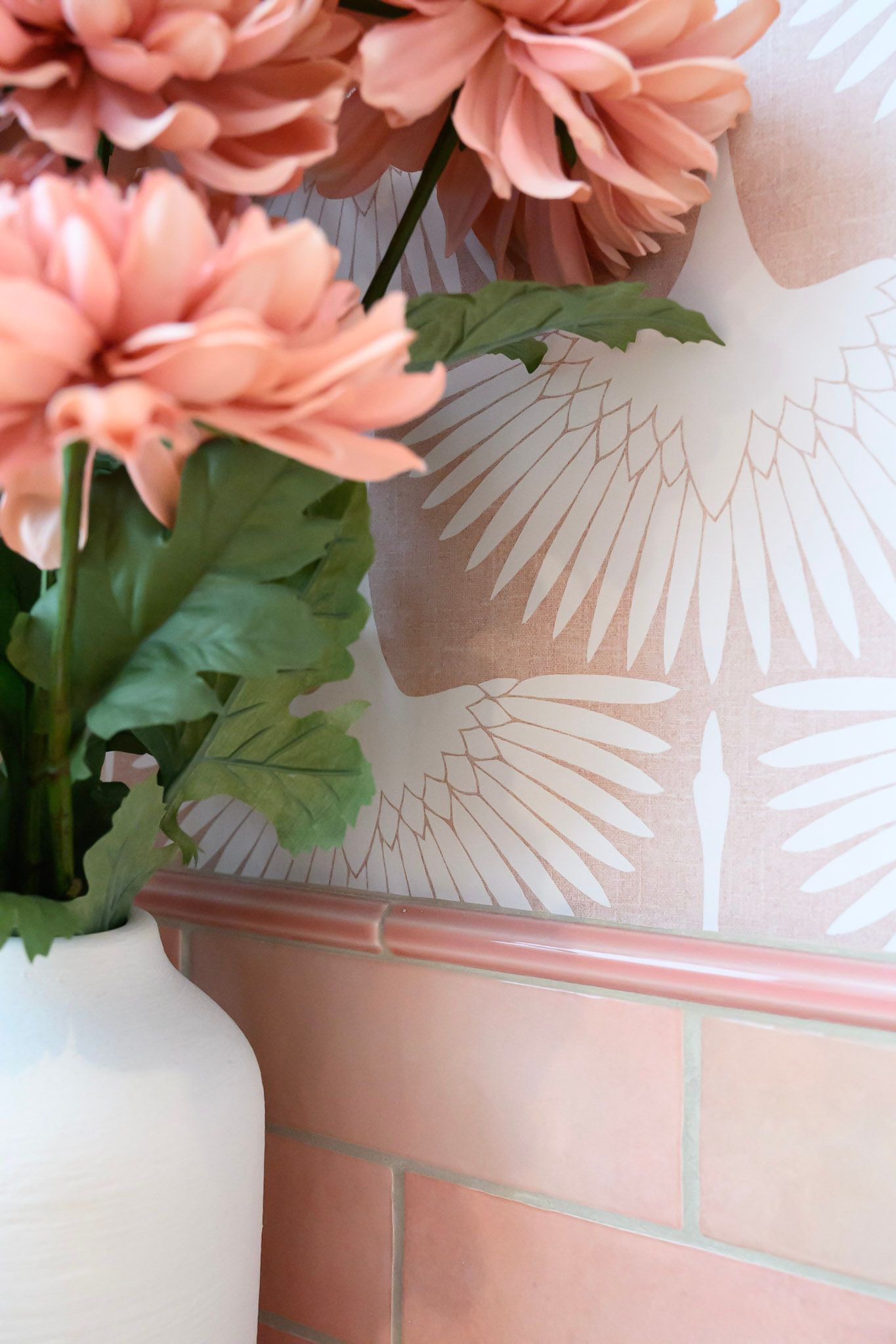 Coral pink artificial flowers in a white vase with geometric pattern against a light background.