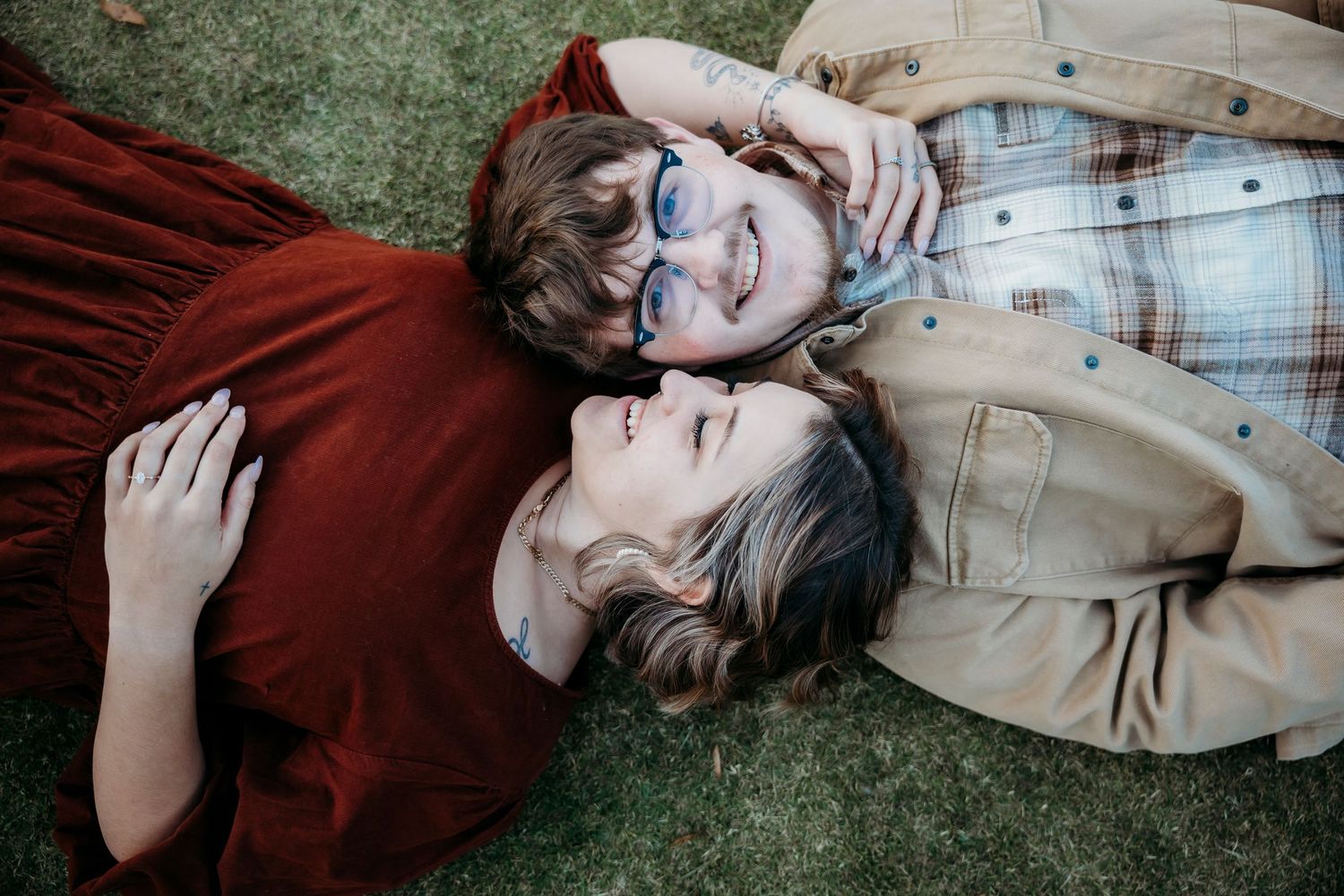 Two people lying on grass in casual autumn attire during engagement photoshoot.