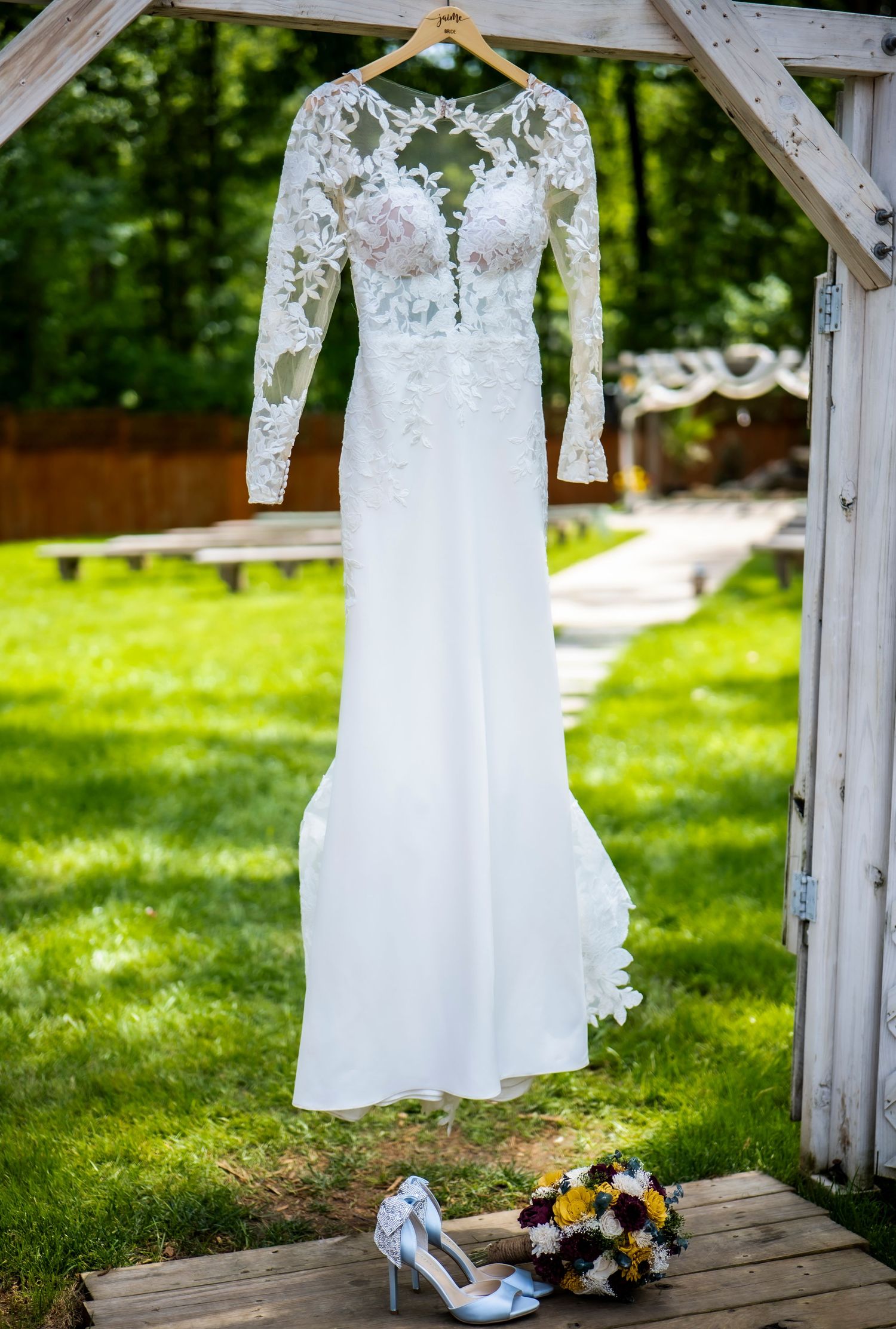 White lace wedding gown displayed on rustic wooden arch with bouquet and shoes below.