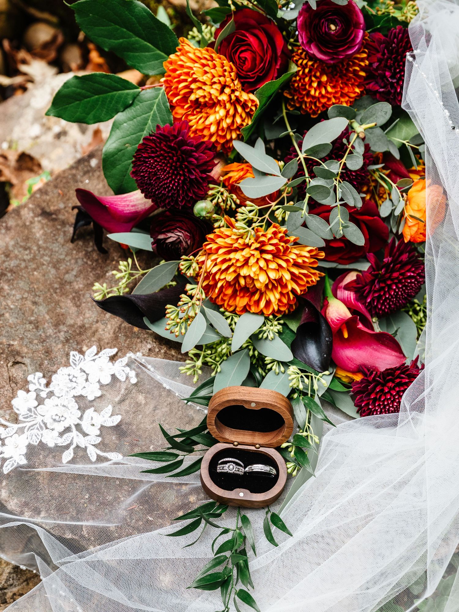 Wedding bouquet with deep red roses, orange dahlias, and eucalyptus leaves against white tulle.