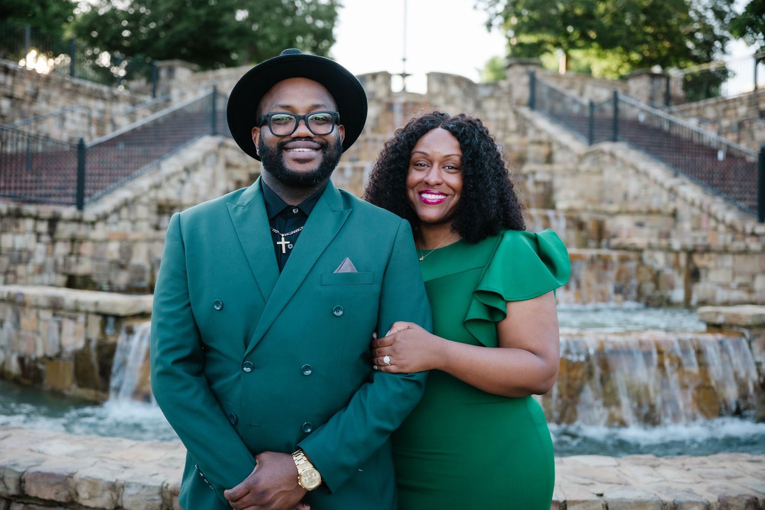 A couple wearing matching green outfits pose together near a stone waterfall feature.