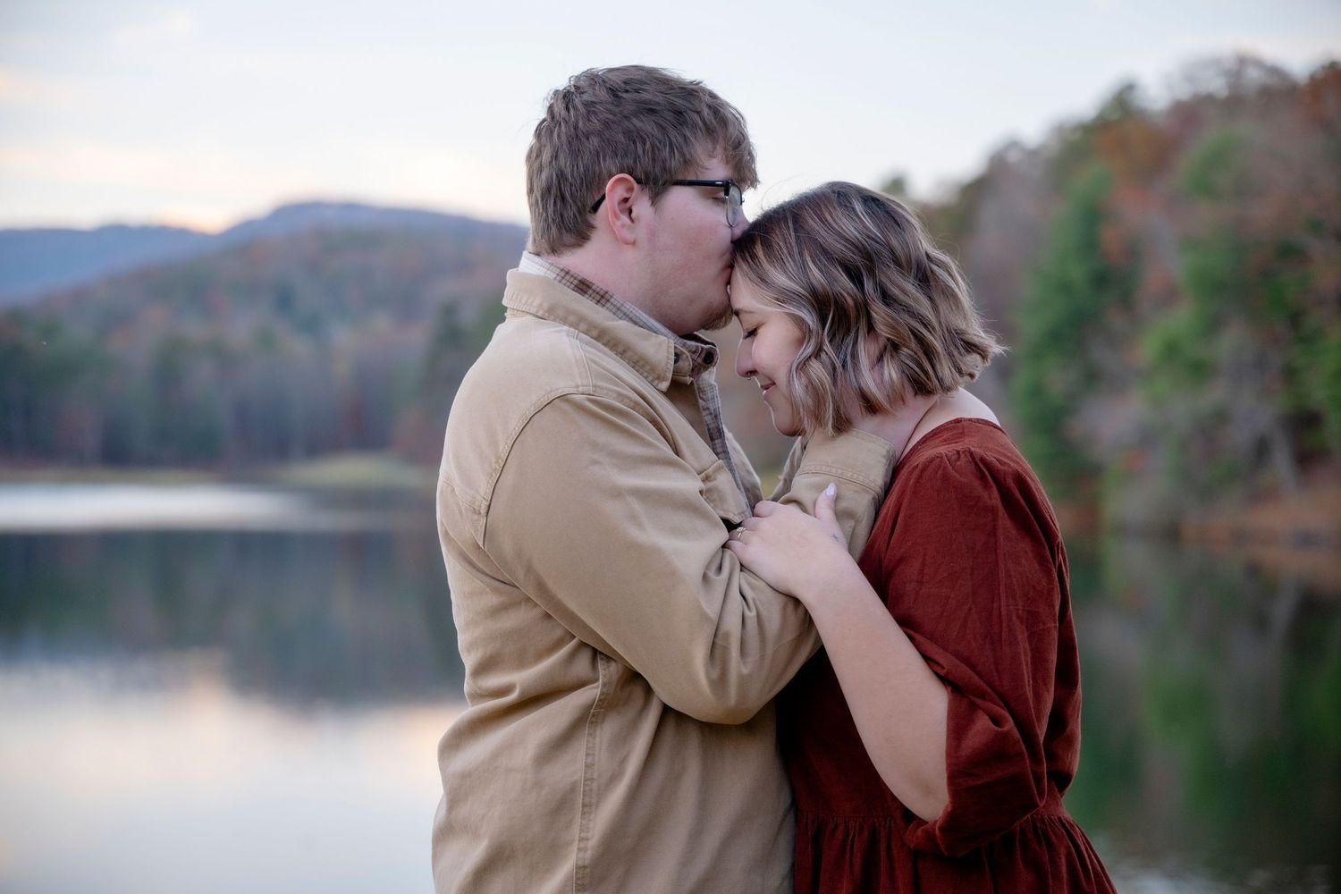 Couple embracing by serene lake with autumn mountains in background.