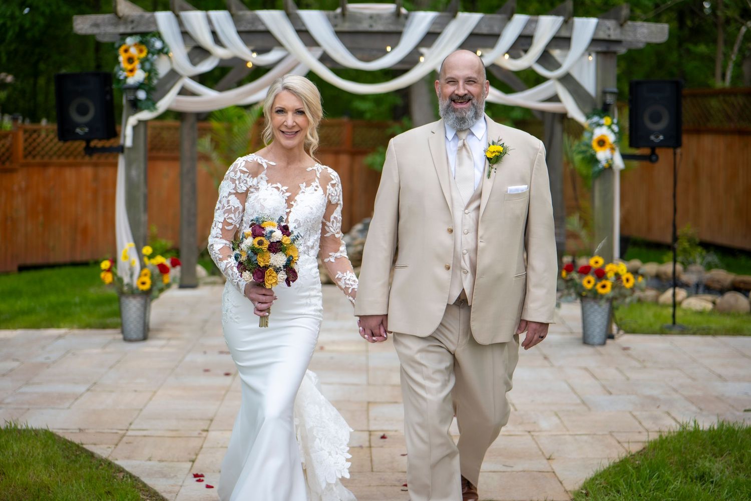 Wedding couple walking together on stone path under white draped arch with yellow floral accents.