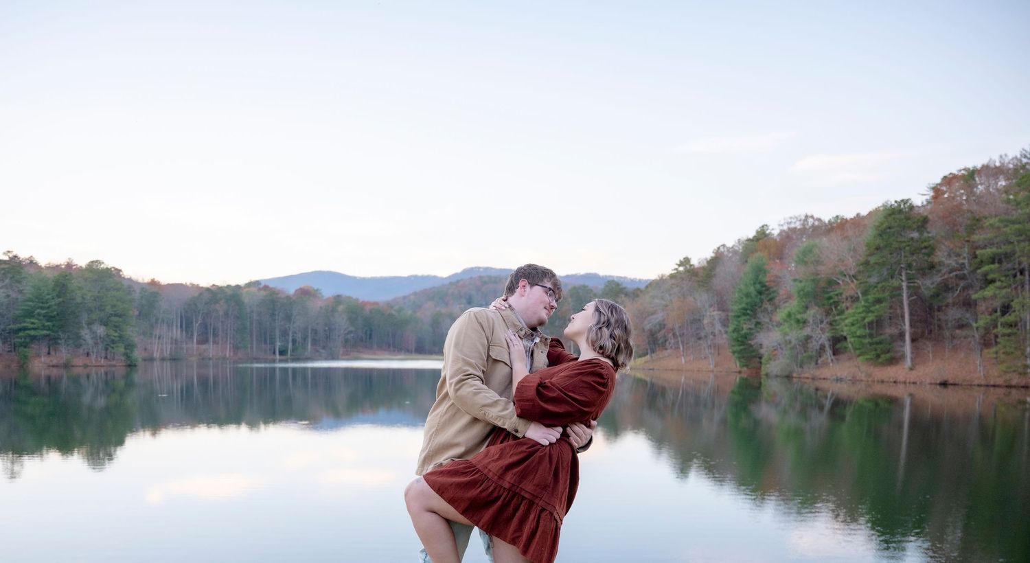 A romantic lakeside scene with mountains and trees reflecting in calm waters at sunset.