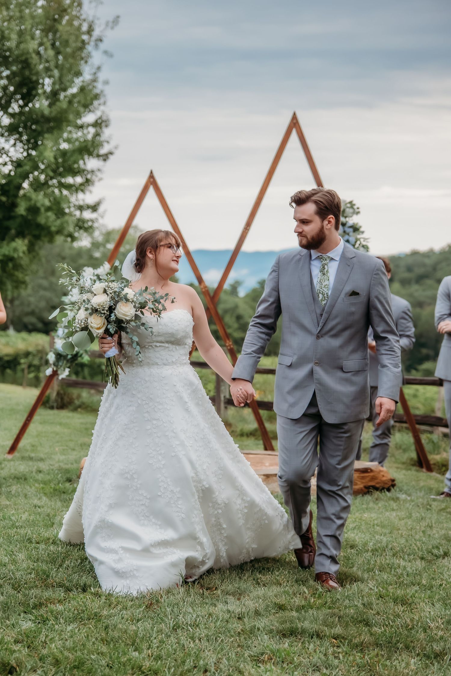 Wedding couple posing near a triangular wooden arch on a grassy hillside with mountain views.