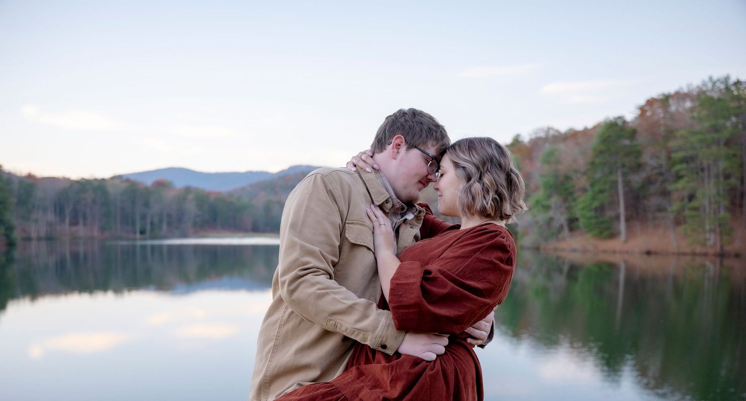 Two people share an intimate moment beside a tranquil mountain lake at dusk.