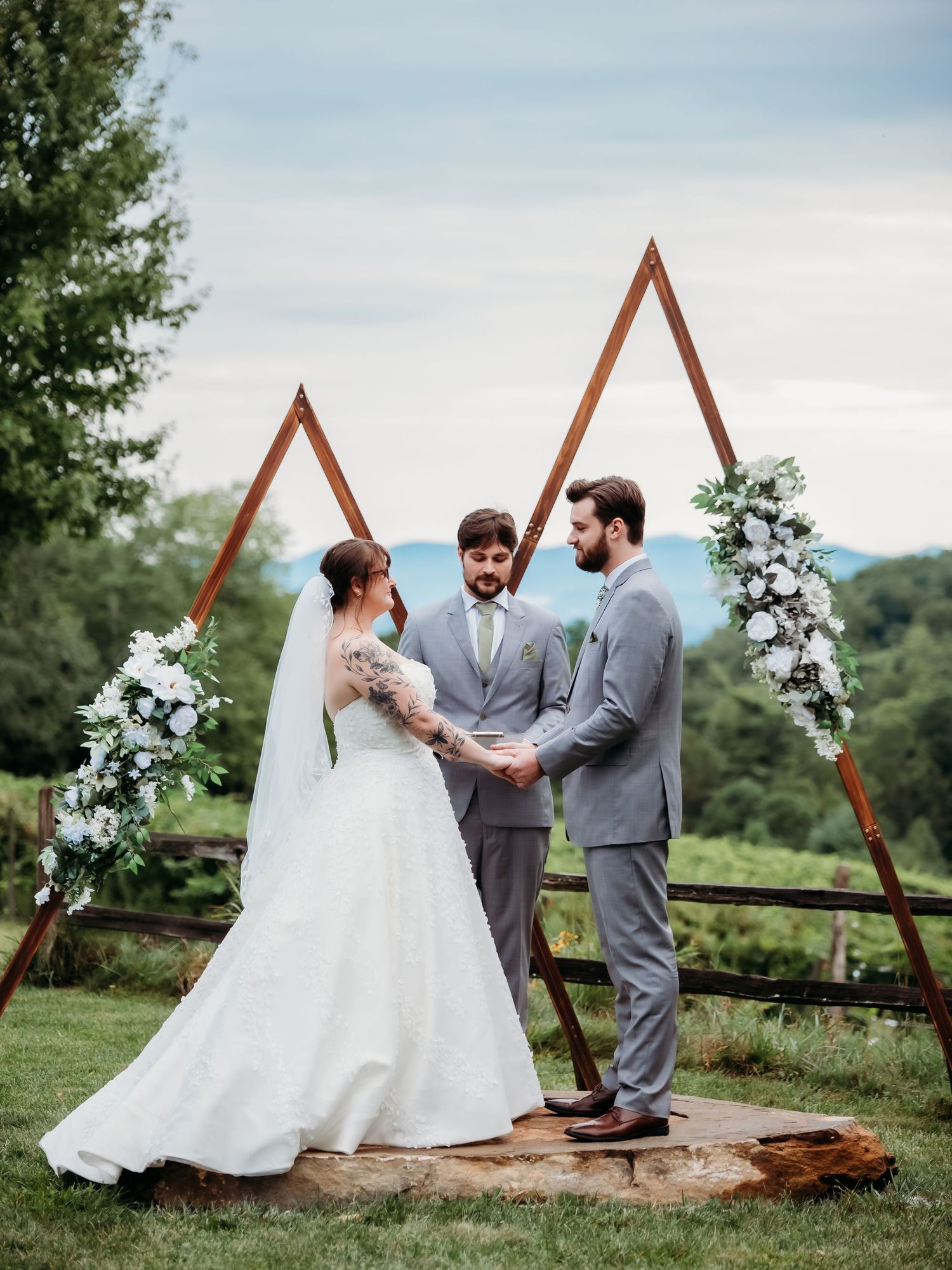 Wedding ceremony setup with a wooden triangle arch decorated with white flowers against a mountain backdrop.