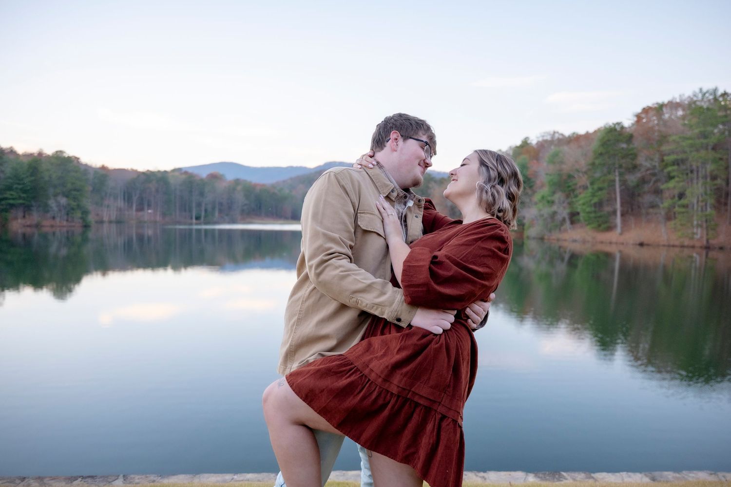 A couple embraces by a serene lake with mountains and autumn trees in the background.