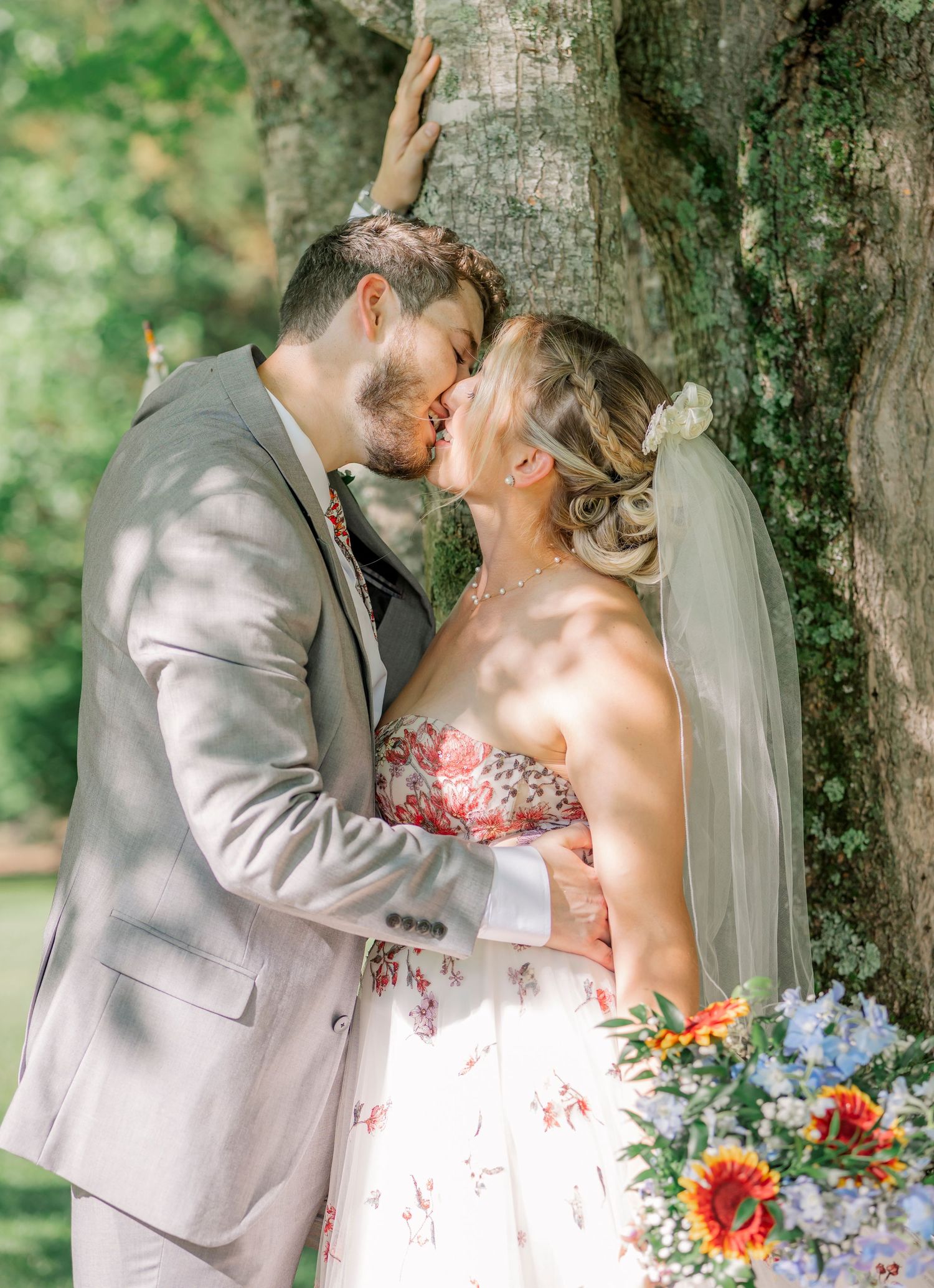 A wedding couple shares intimate moments under a large tree in a sunny garden setting.