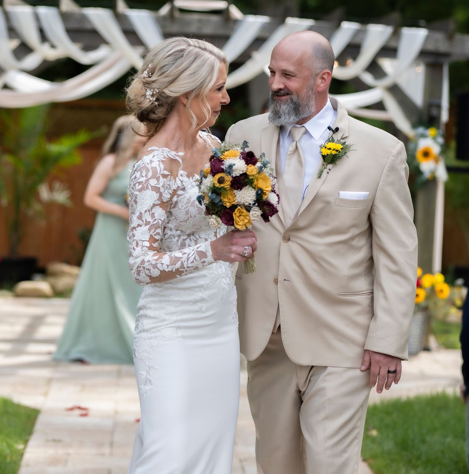 A romantic wedding moment featuring a couple in a tan suit and lace gown with sunflower bouquet under white decor.