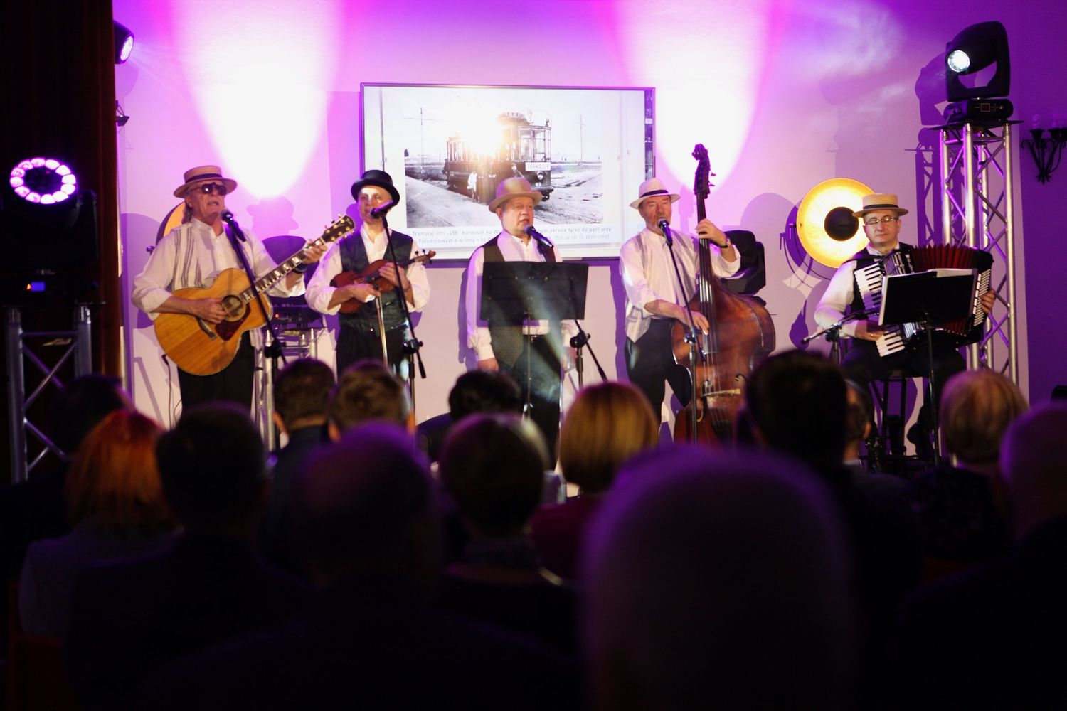 Five-piece band performing live on stage with string instruments at evening event.