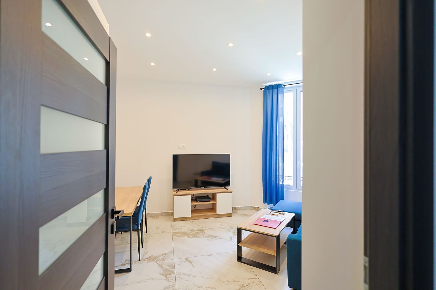 Modern apartment living room with flat-screen TV, blue curtains, marble floors, and minimalist furniture in a bright white space.