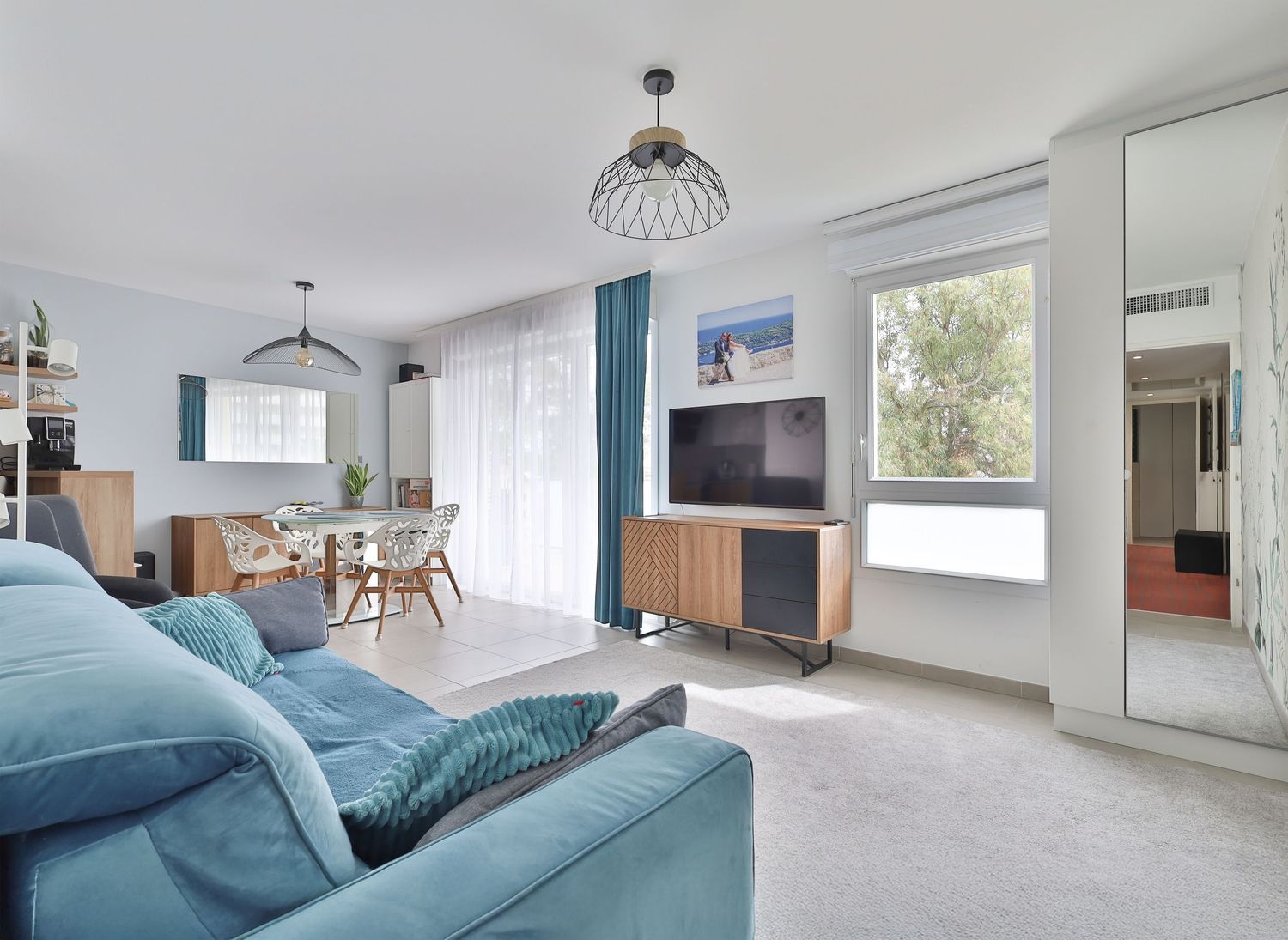 Bright apartment living room featuring blue sofa, wooden TV unit, and dining area with natural light.
