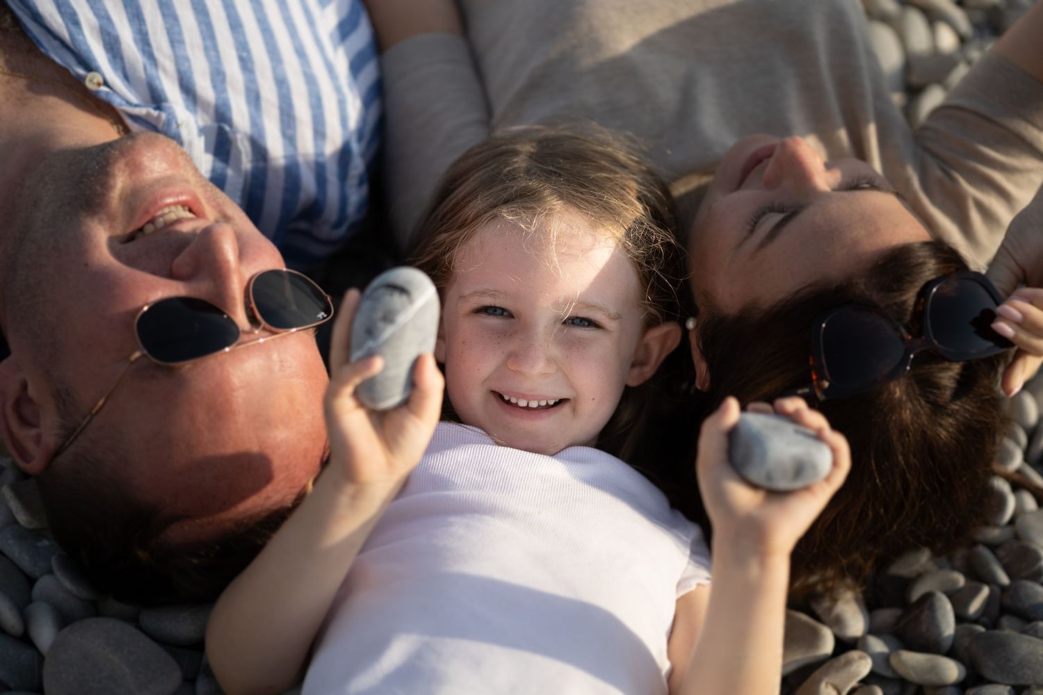 Cheerful young girl lying between her parents on a pebble beach, all holding smooth stones and smiling at the camera.