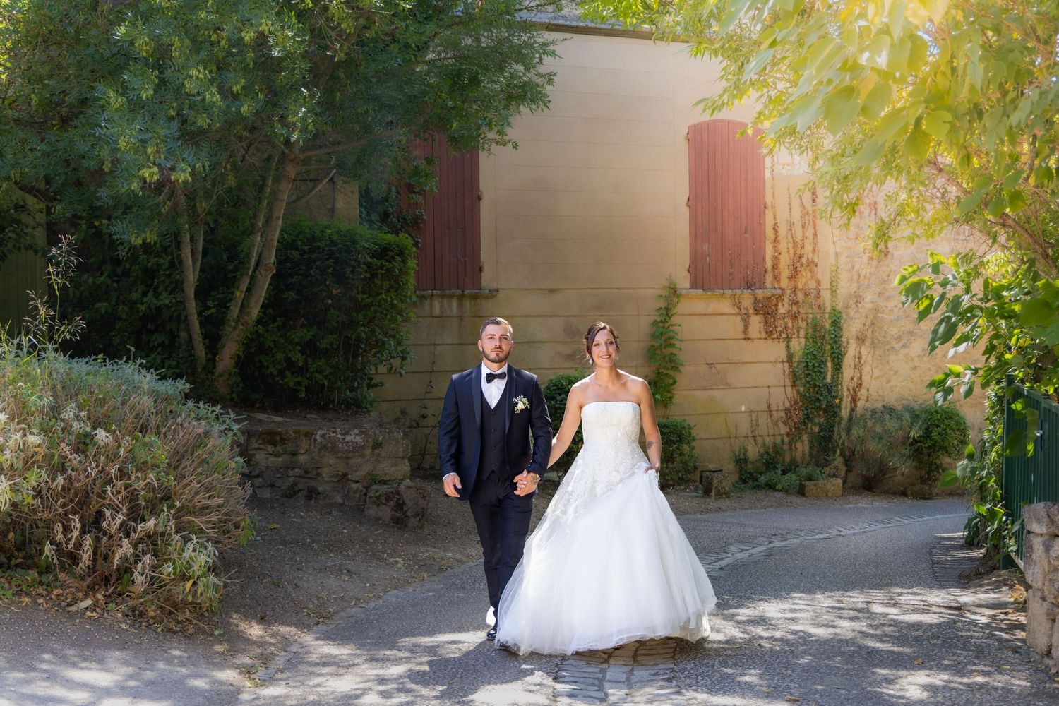 Bride and groom walking hand in hand outdoors near a rustic building surrounded by lush green trees on their wedding day.