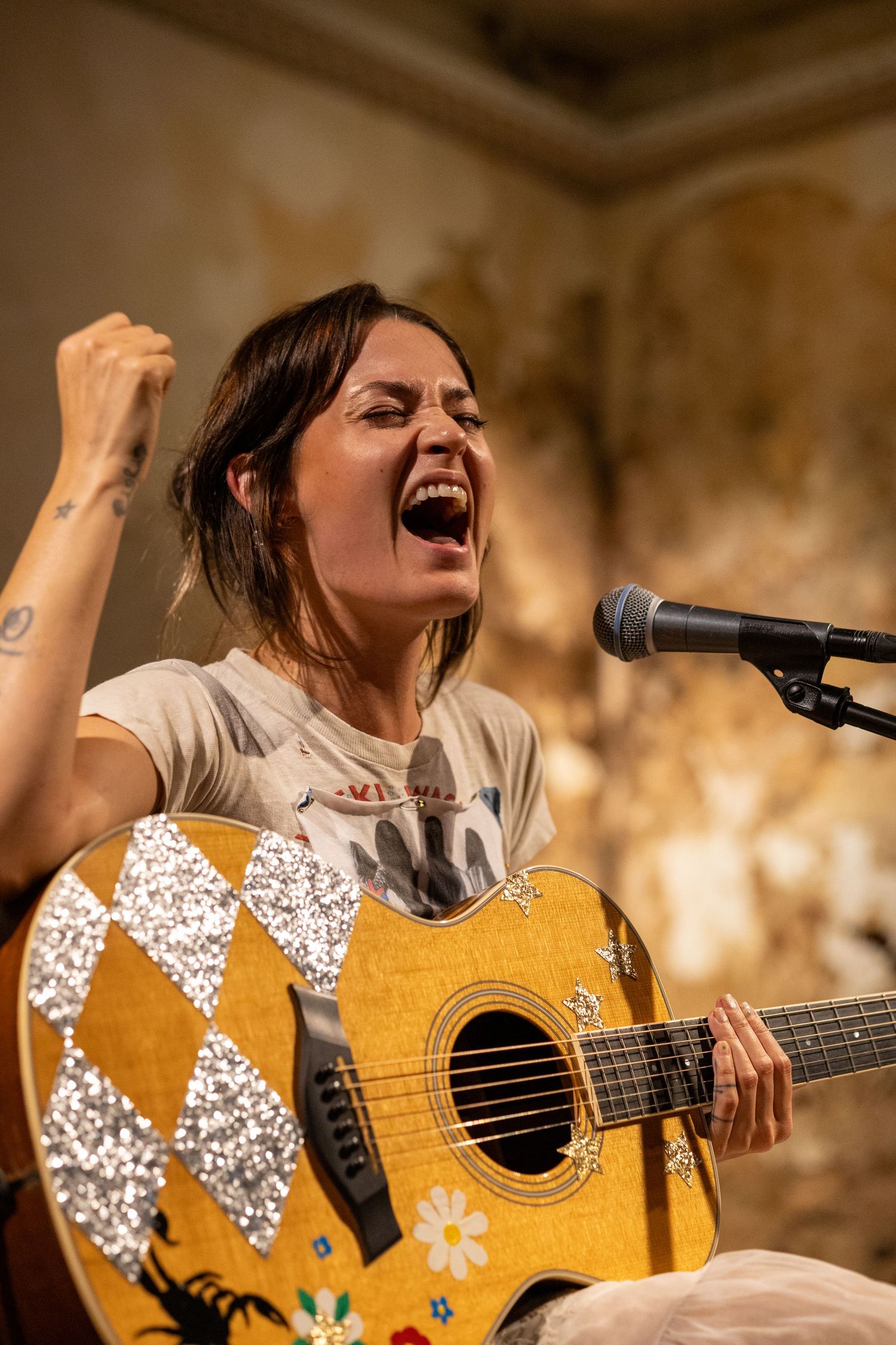 Woman passionately singing and playing decorated acoustic guitar at microphone stand.