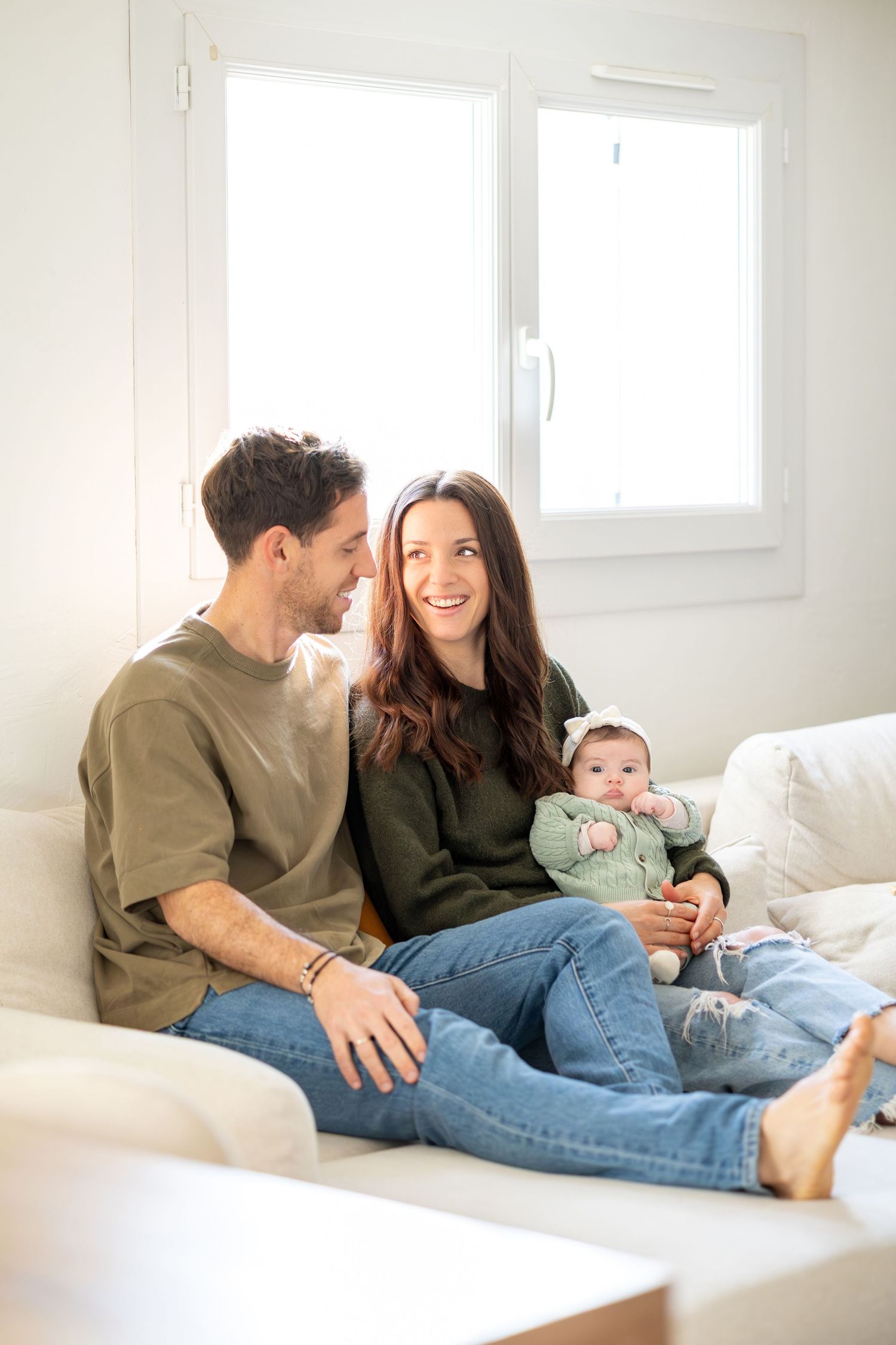 Happy young couple sitting on white sofa smiling at each other while holding their baby.