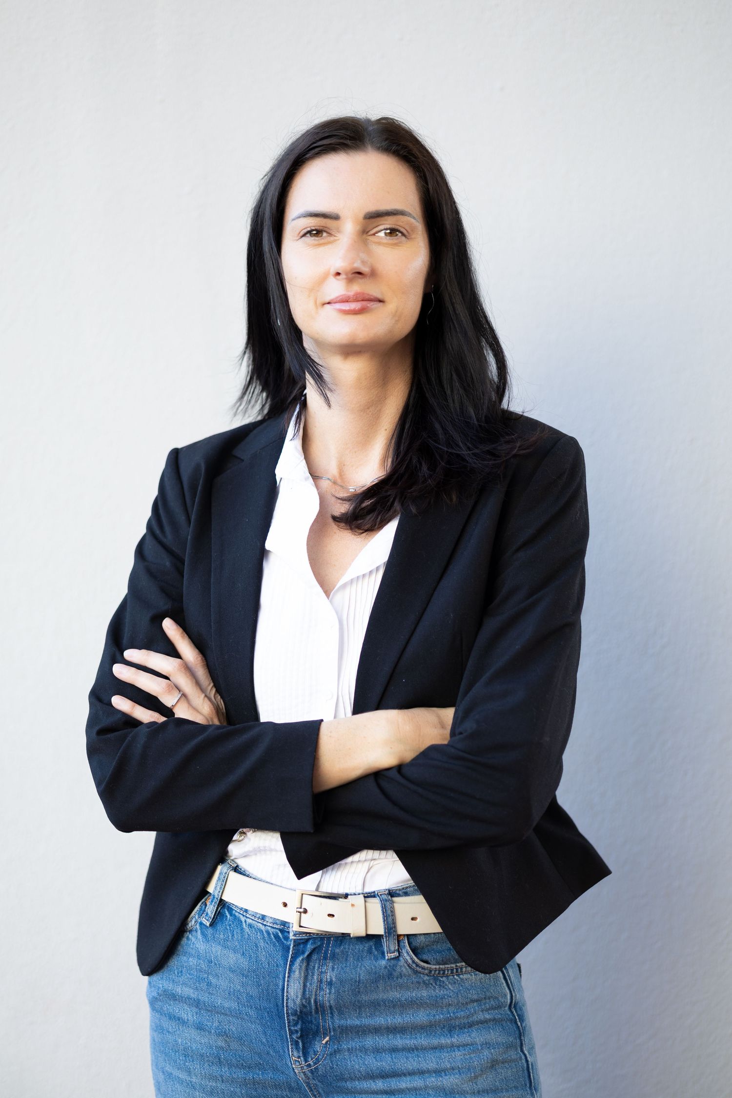 Professional woman in black blazer and jeans standing with arms crossed against a light gray background.