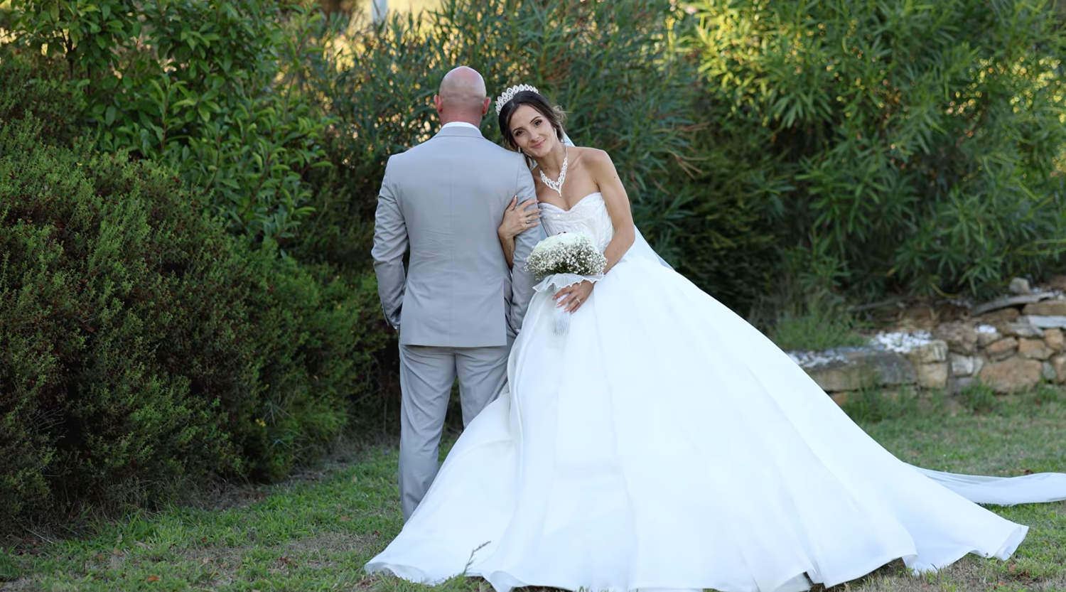 Bride in white ball gown and groom in gray suit pose together outdoors surrounded by lush green trees on their wedding day.