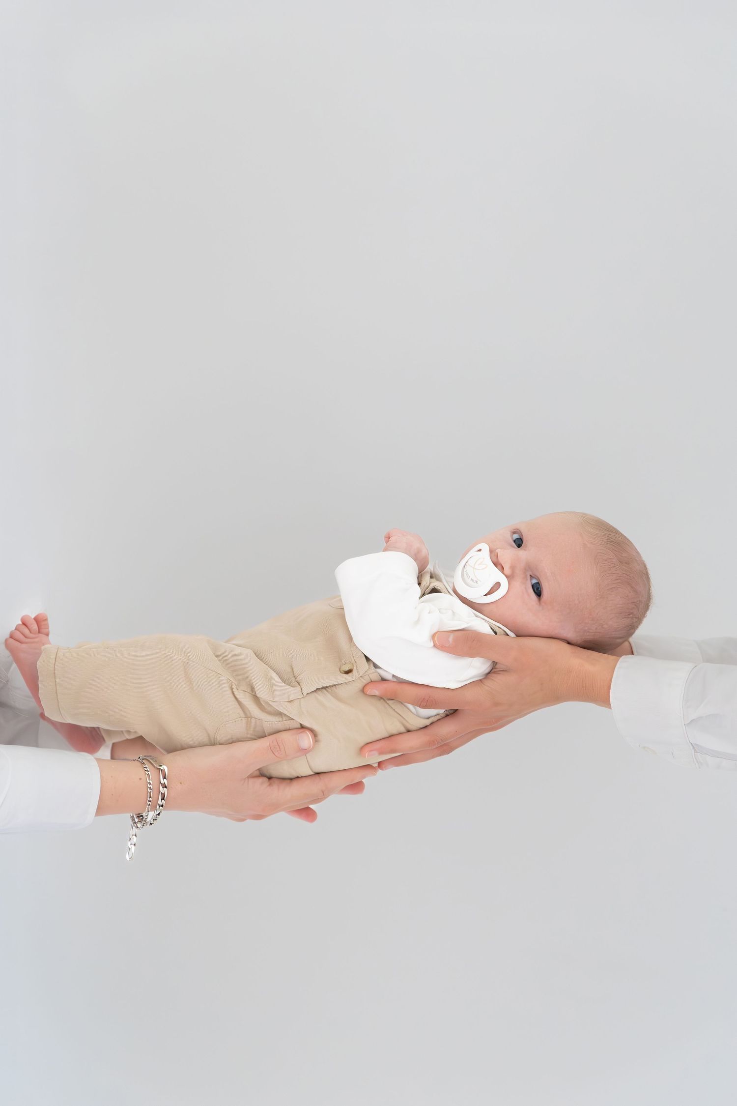 Two hands carefully holding a newborn baby with a pacifier against white background.