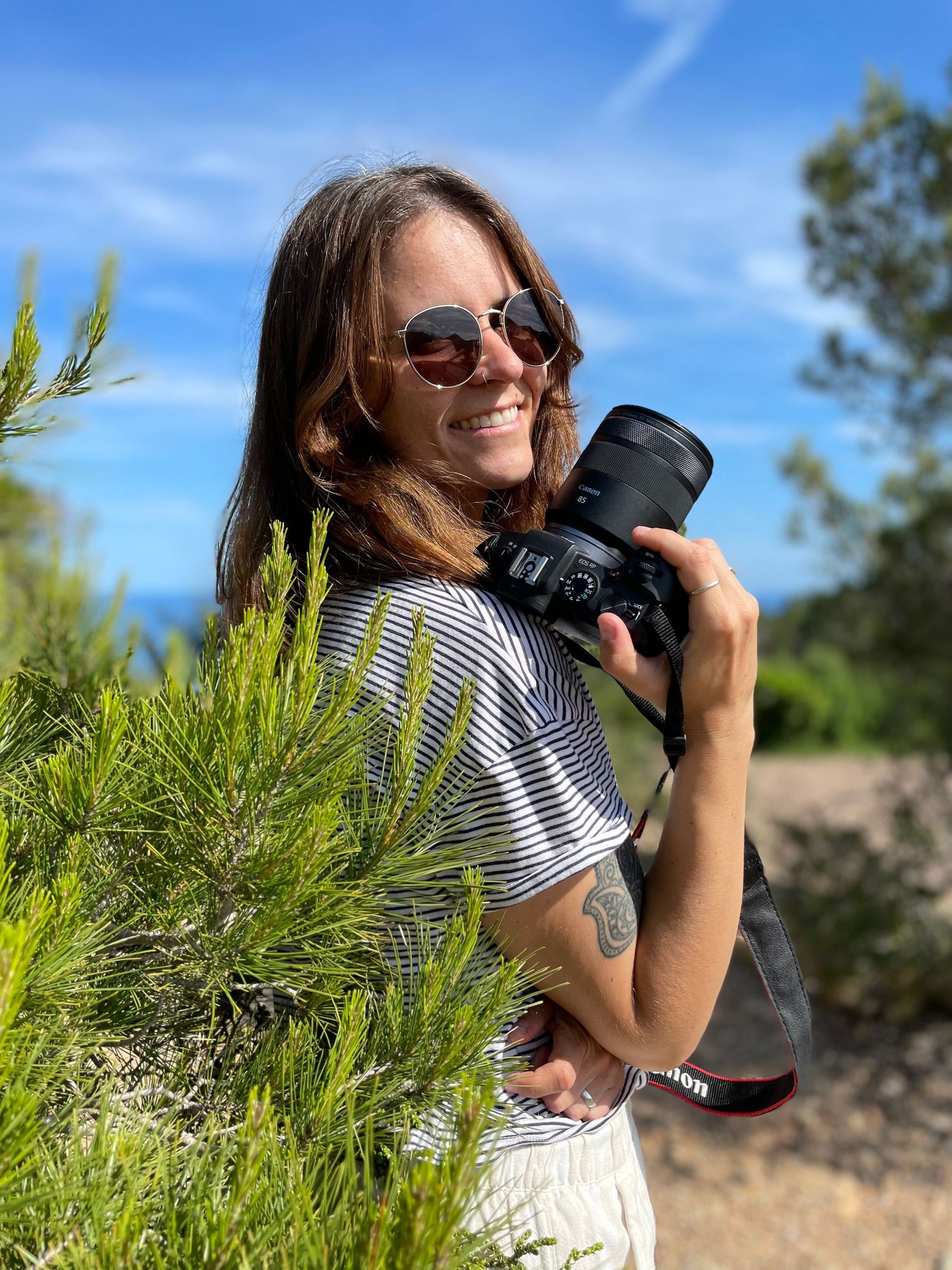 Smiling woman photographer holding a Canon camera outdoors among green plants under a blue sky.