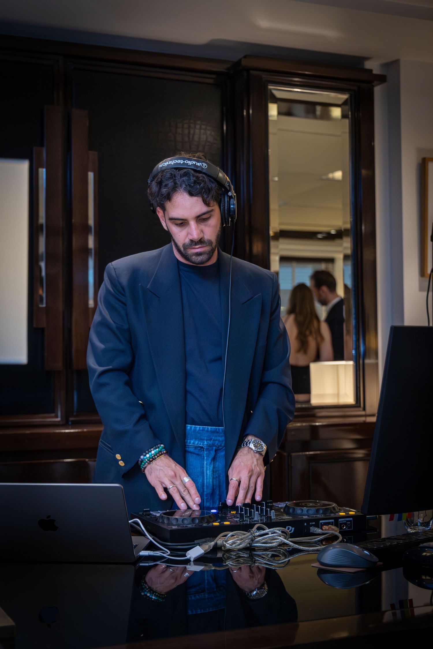 A DJ wearing headphones and a dark blazer operates a mixer and laptop at an indoor event.