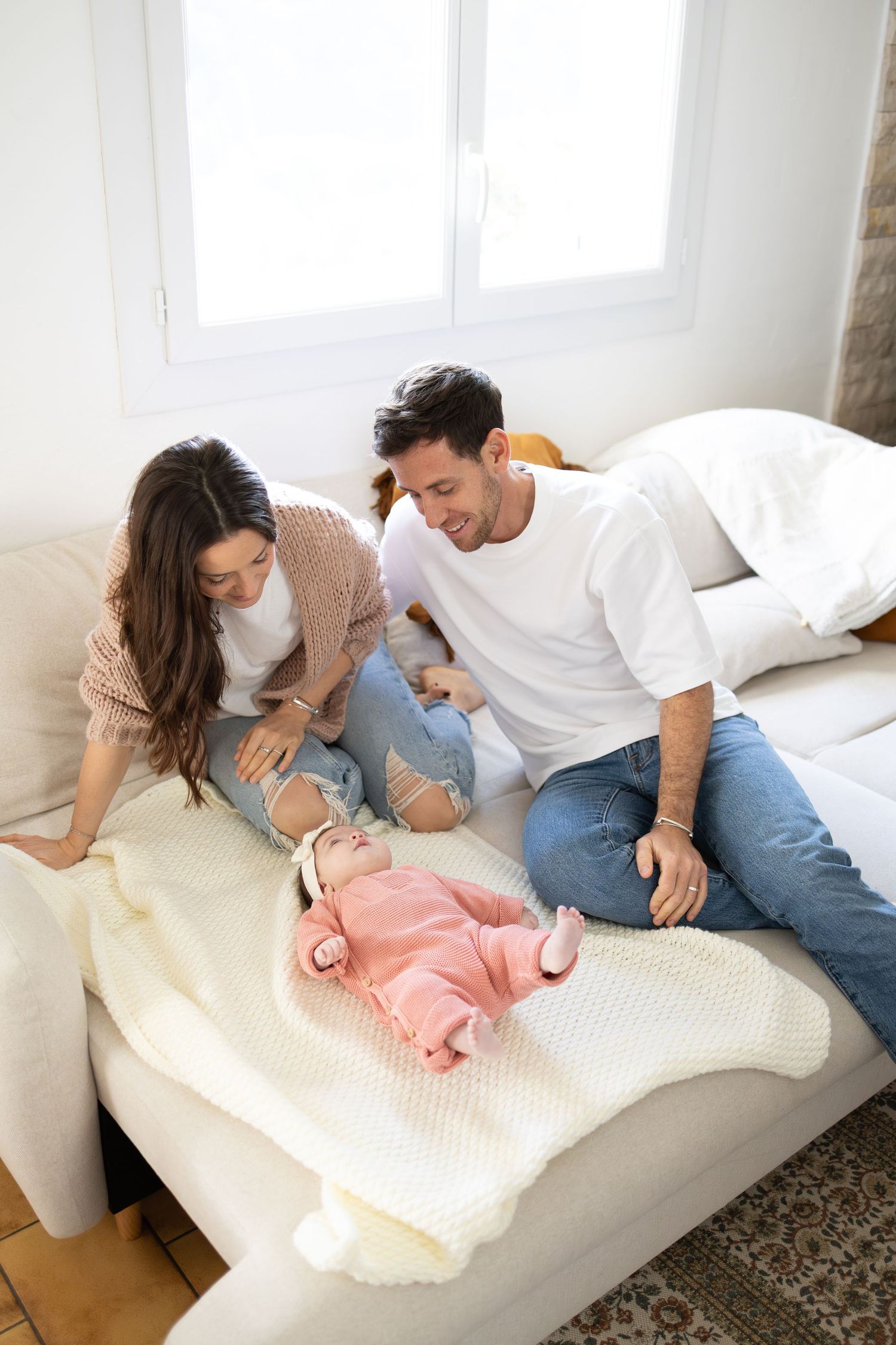 Young couple smiling and bonding with their newborn baby lying on white blanket on sofa.
