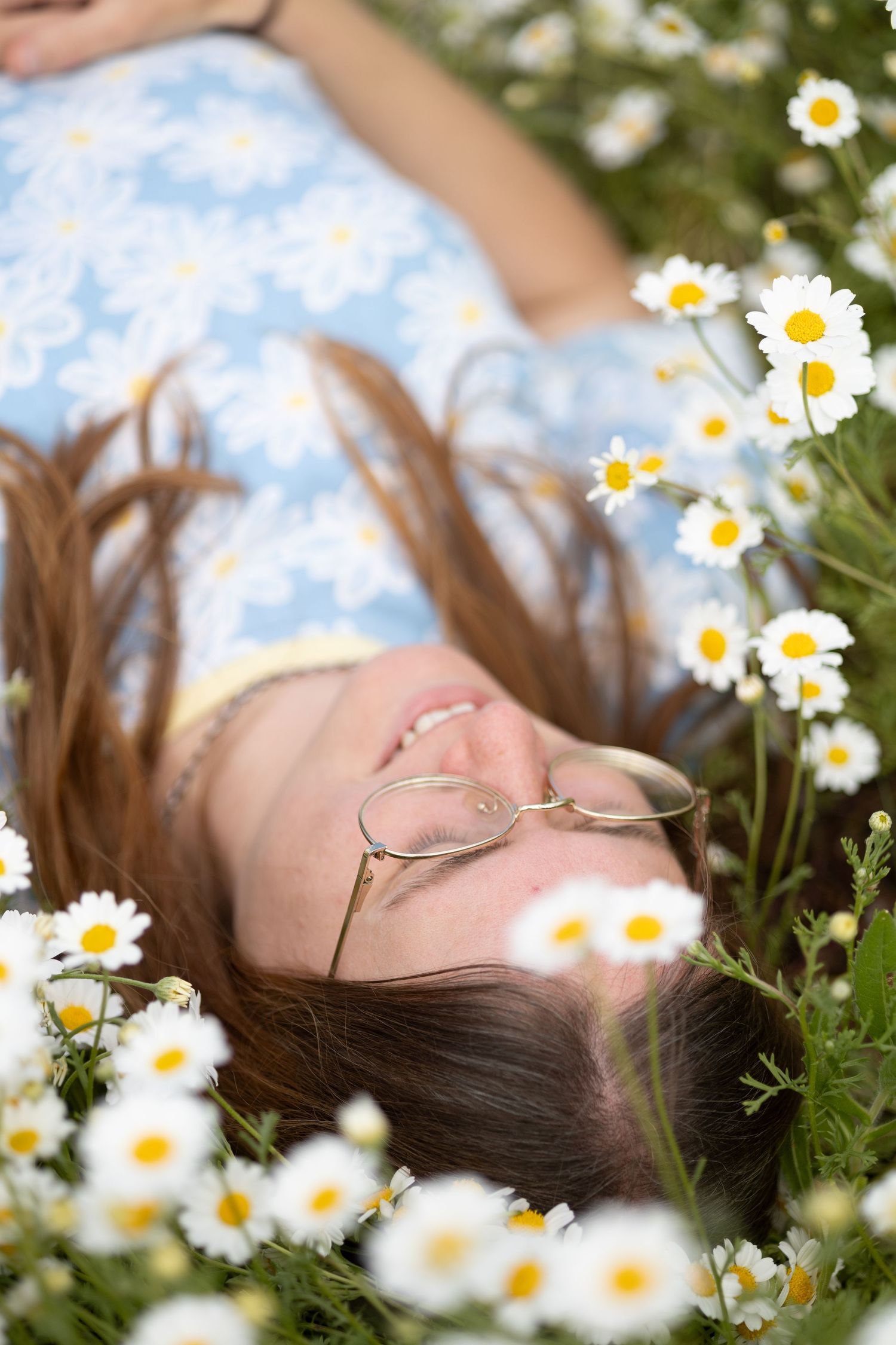 Young woman with glasses lying in a field of white daisies wearing a blue floral dress.